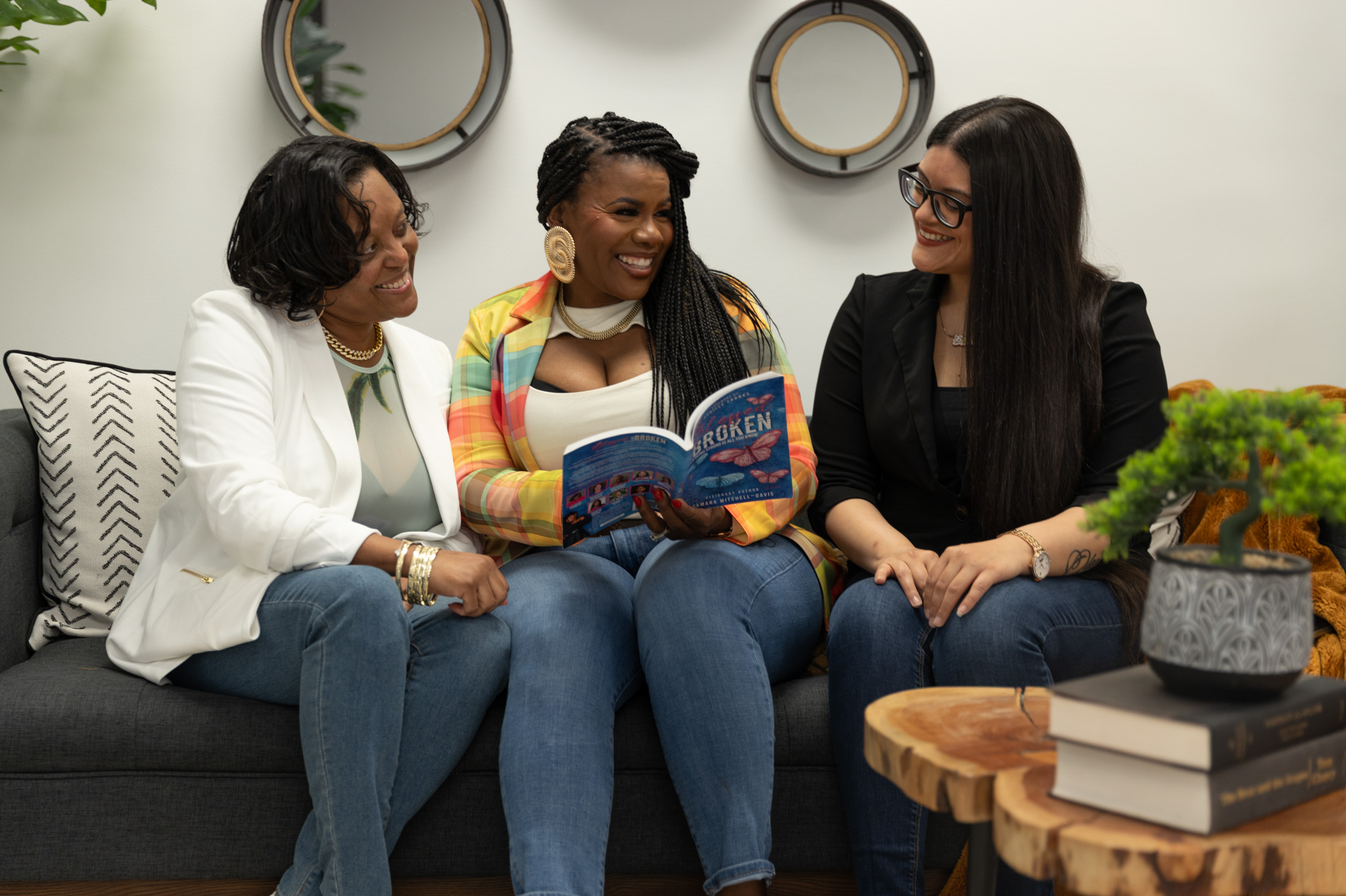 Three women sitting on a sofa, smiling and talking, with books and decorative items in the background.