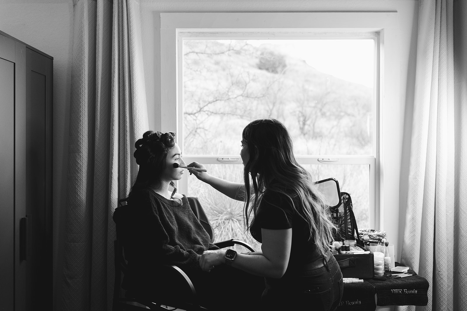 Two women sit facing each other near a window, one applying makeup to the other, in a room with curtains and a table.