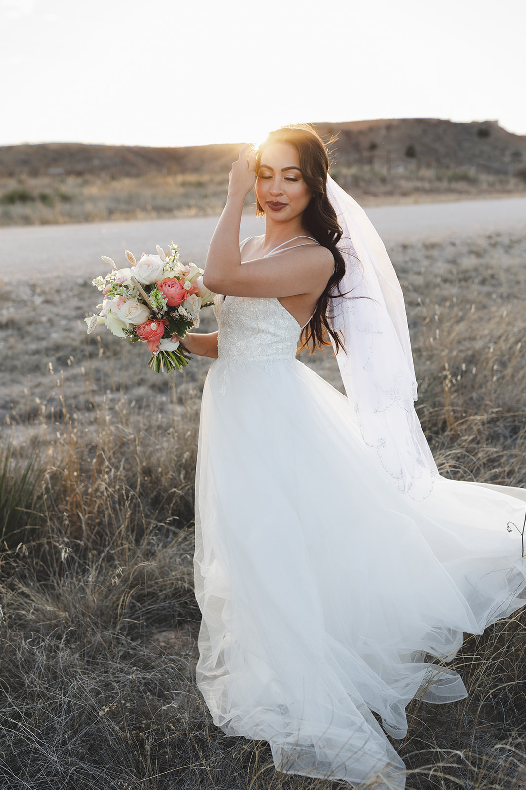 Bride in wedding dress holding a bouquet outdoors, with her hand on her head and eyes closed.