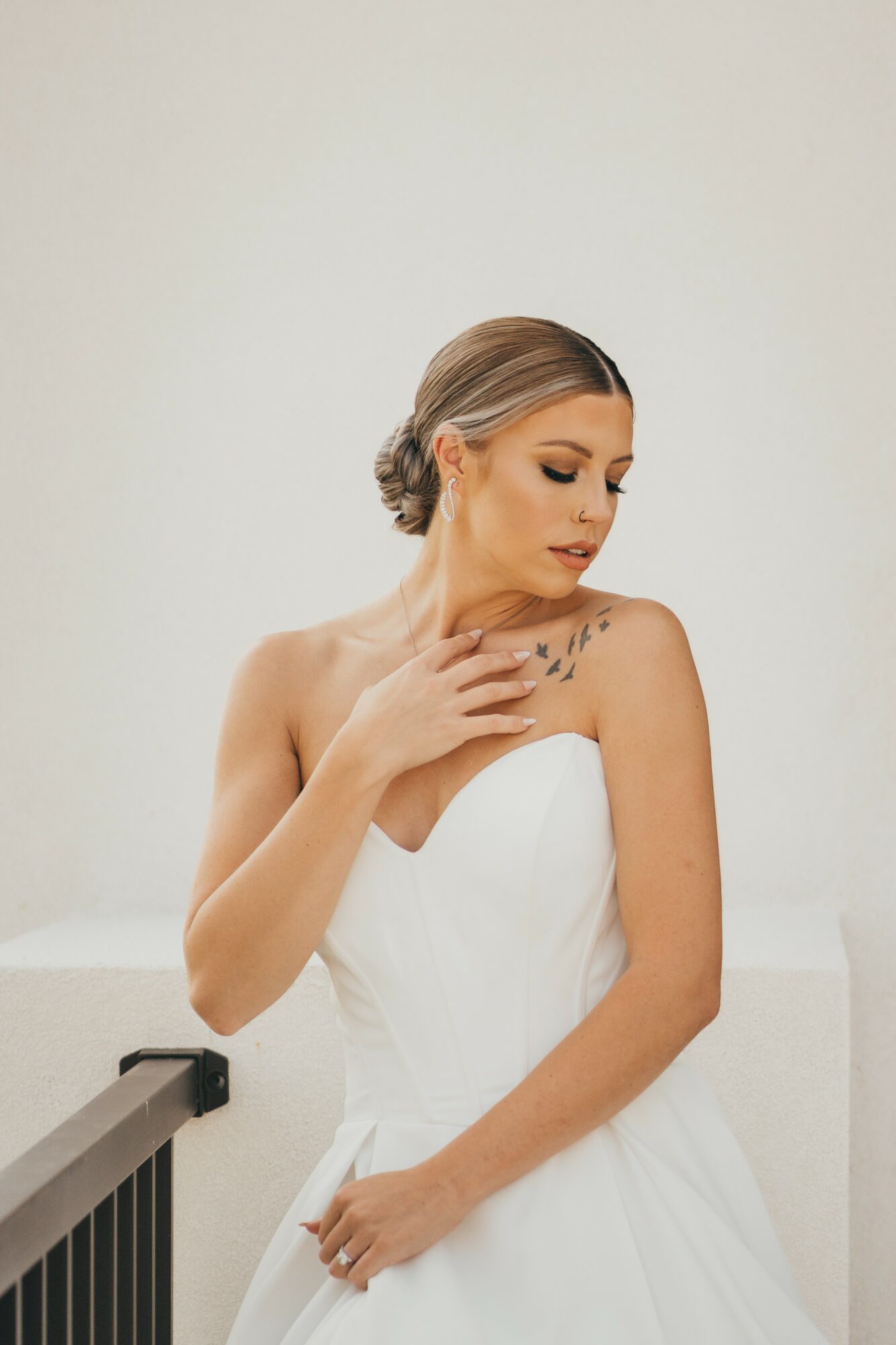 Woman in a white strapless dress with her hand on her chest, looking down, against a plain background.