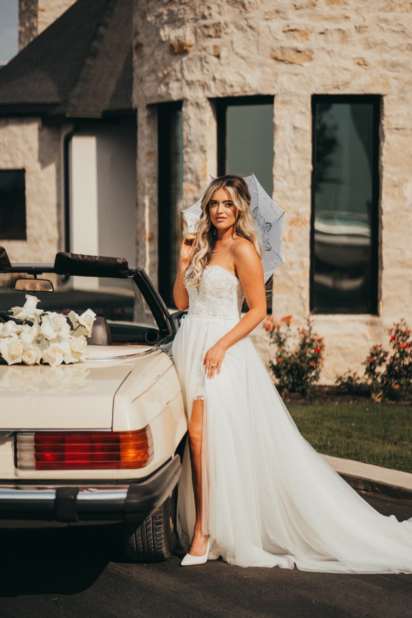 Bride in wedding dress standing next to a car with flowers, in front of a stone building.