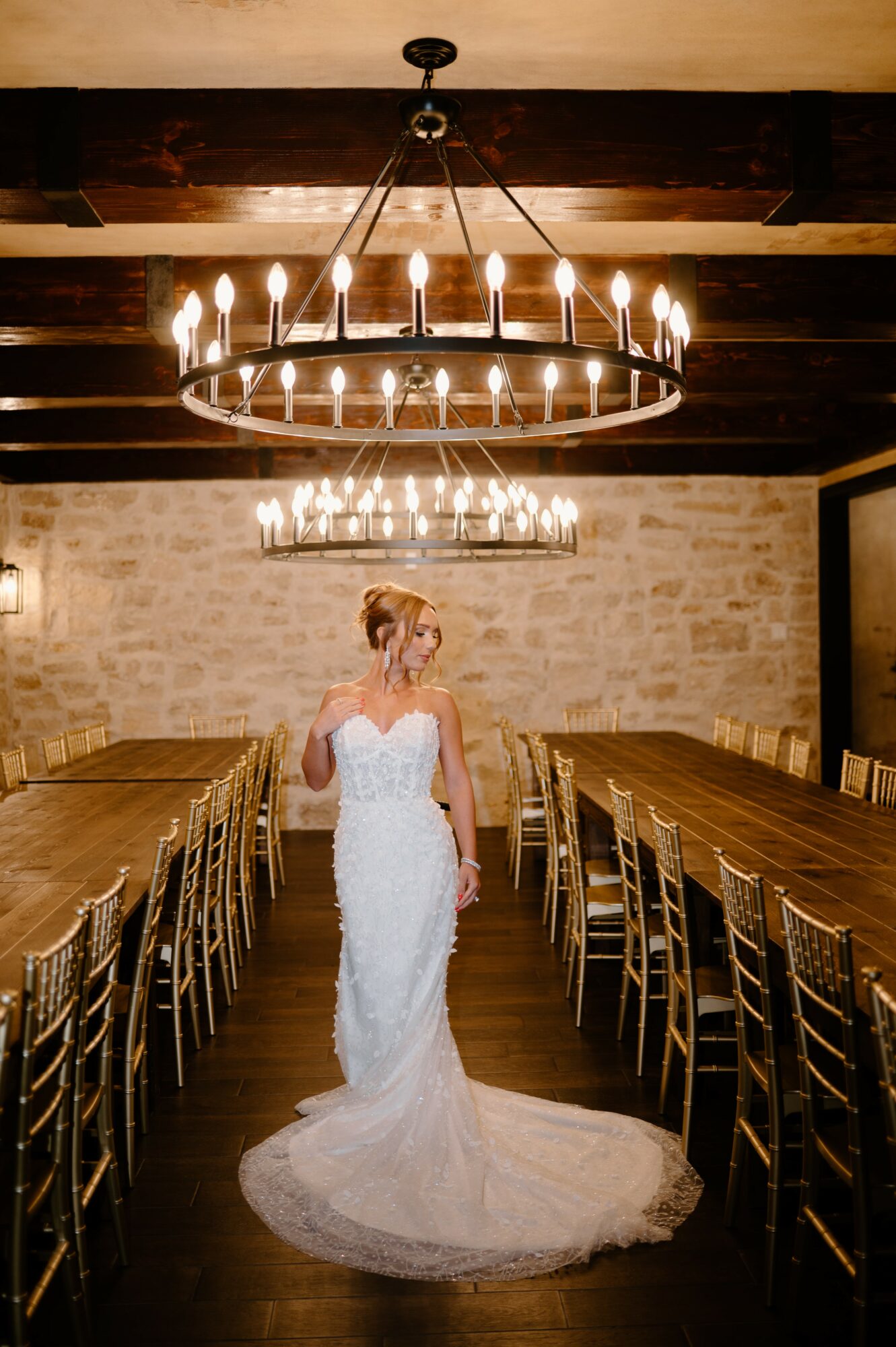 Woman in wedding dress walking down a long dining table in a rustic room with chandeliers.