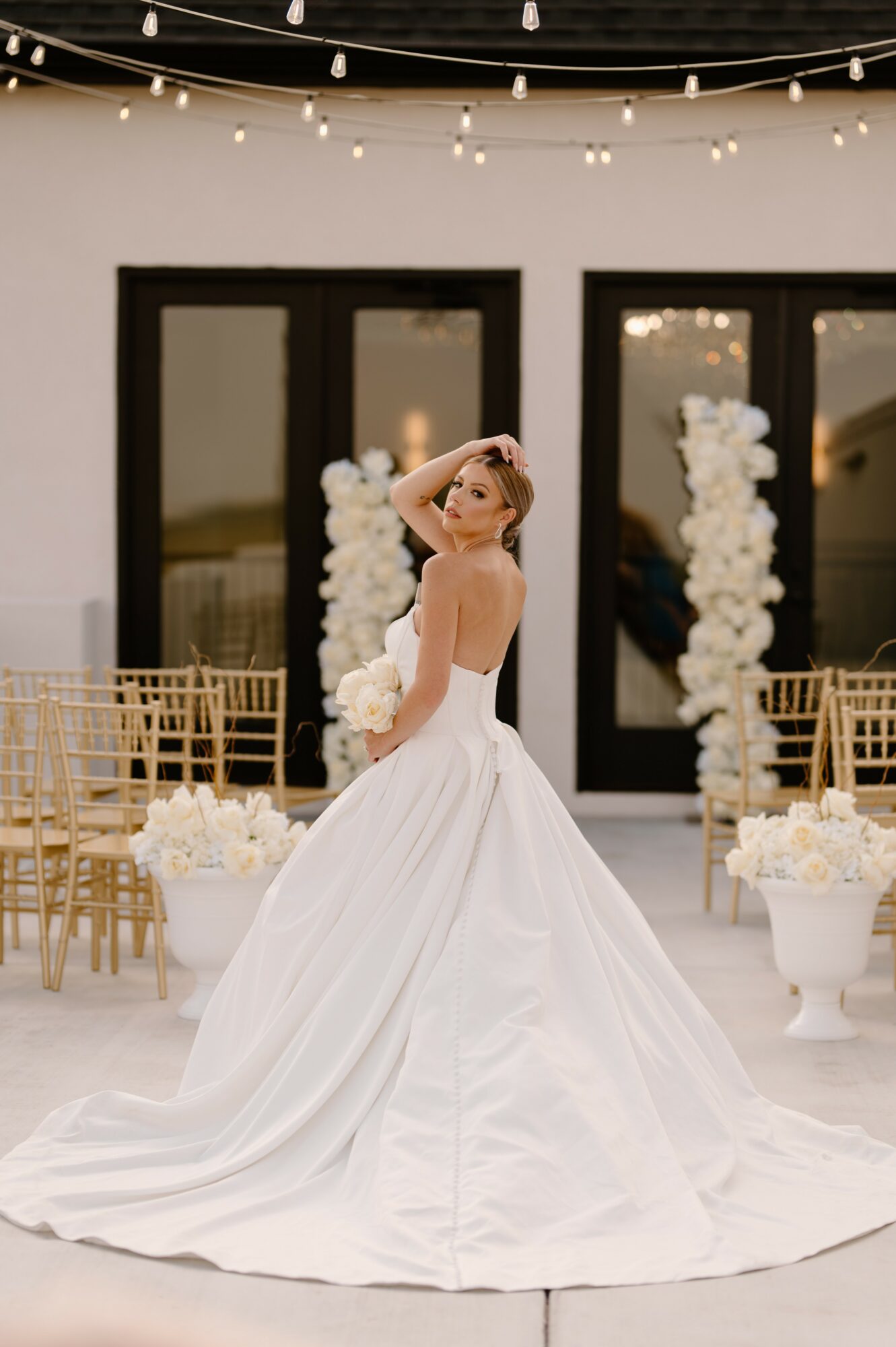 Bride in a white wedding gown standing indoors with floral decorations and chairs, looking at the camera, hand on head.