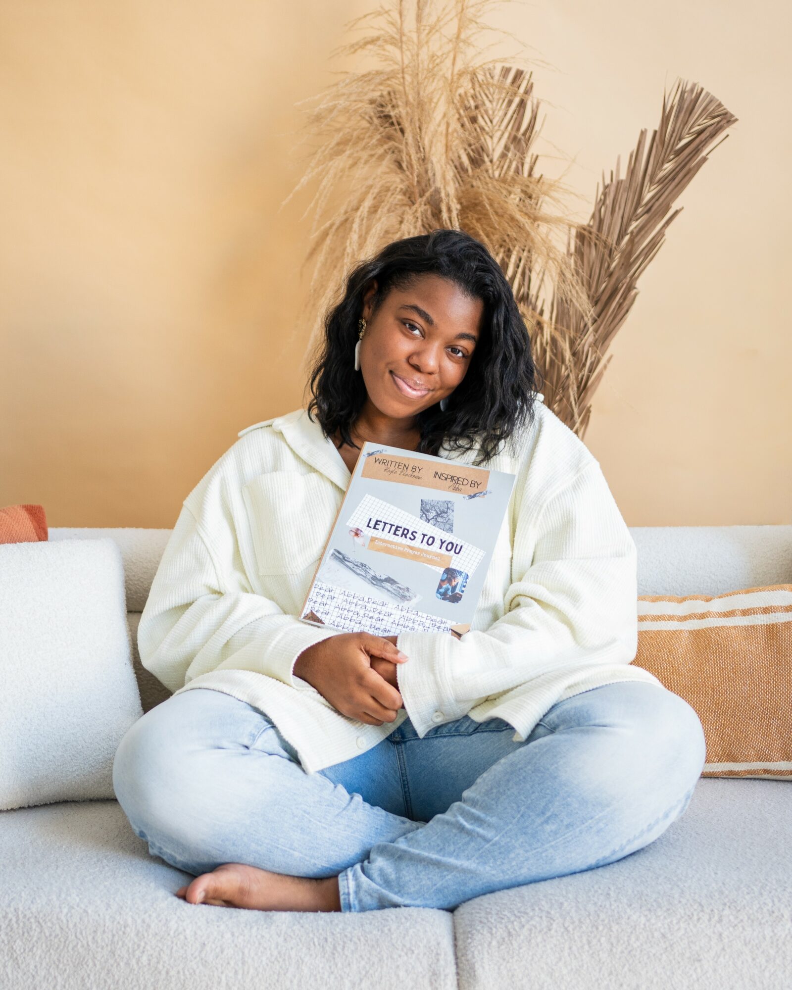 Smiling woman sitting cross-legged on a sofa holding a letter, with dried plants behind her, in a cozy room.