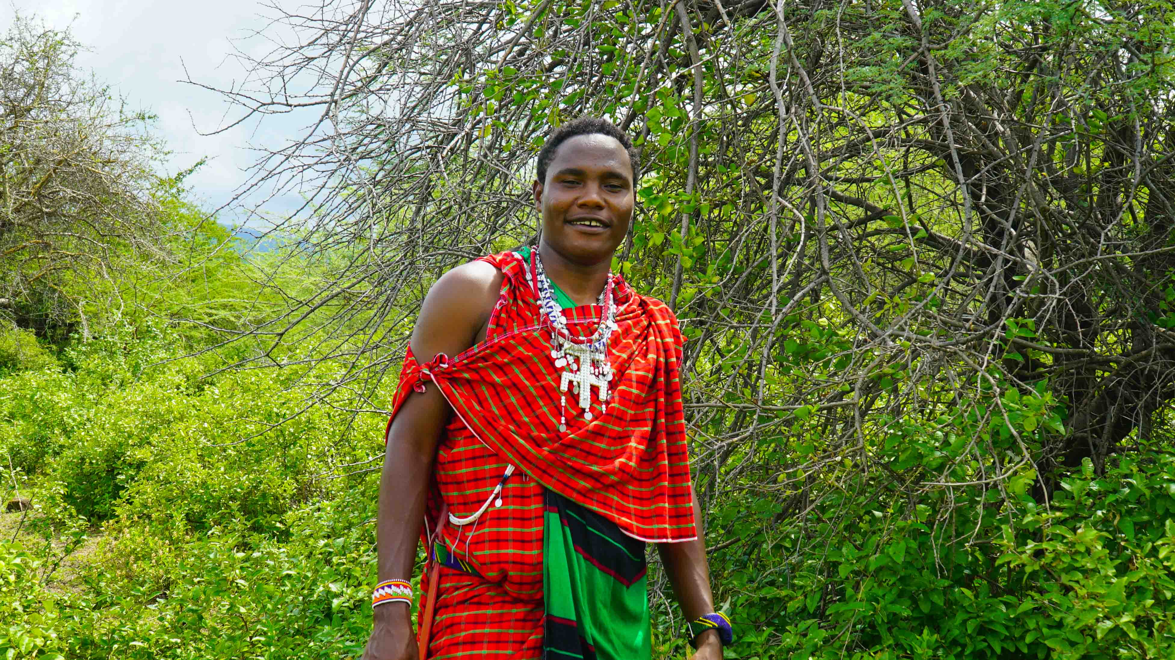 Person standing outdoors among green trees, wearing traditional red and green clothing, smiling.