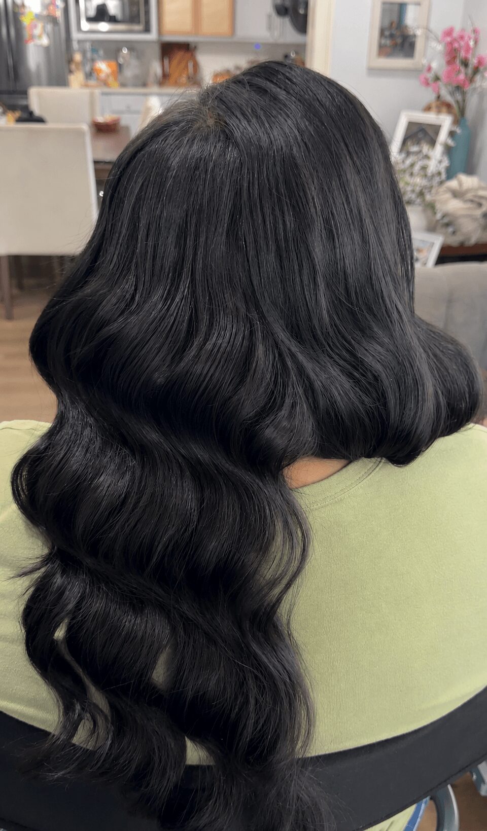 Back of a person with long, wavy black hair, sitting in a room with framed pictures and flowers in the background.