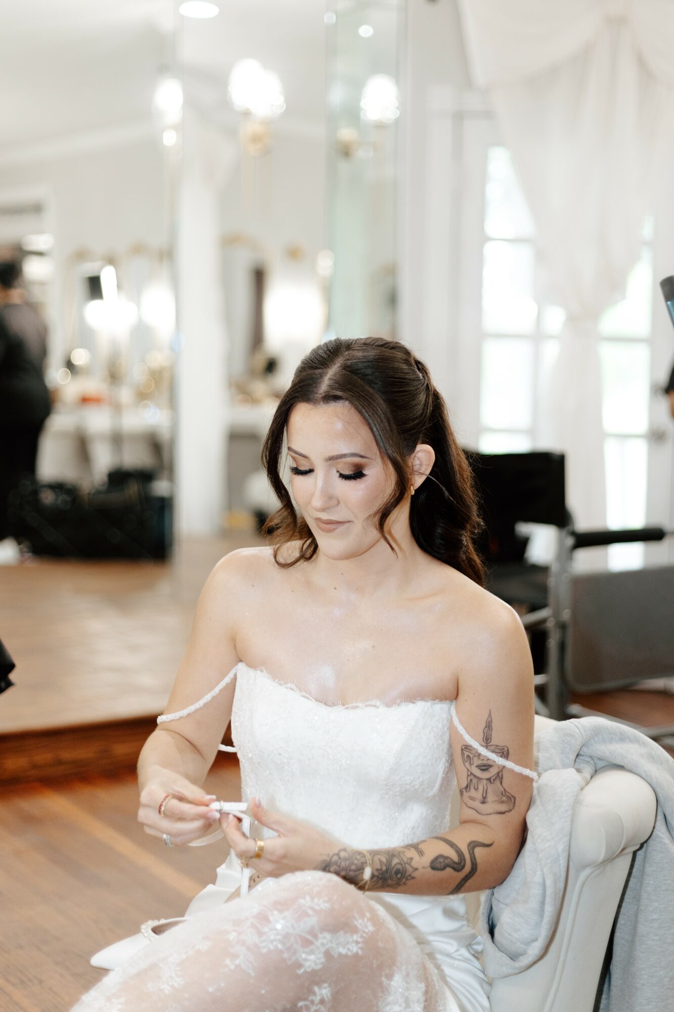 Woman in a white strapless dress sitting on a chair, looking at her phone in a bright room with large windows.