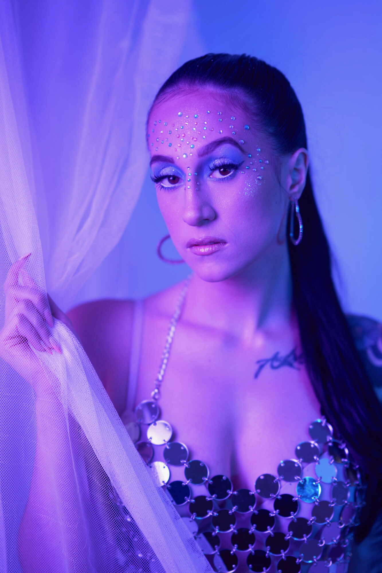 Woman with makeup and glitter on face, wearing a shiny top, holding sheer fabric, in a purple-lit setting.
