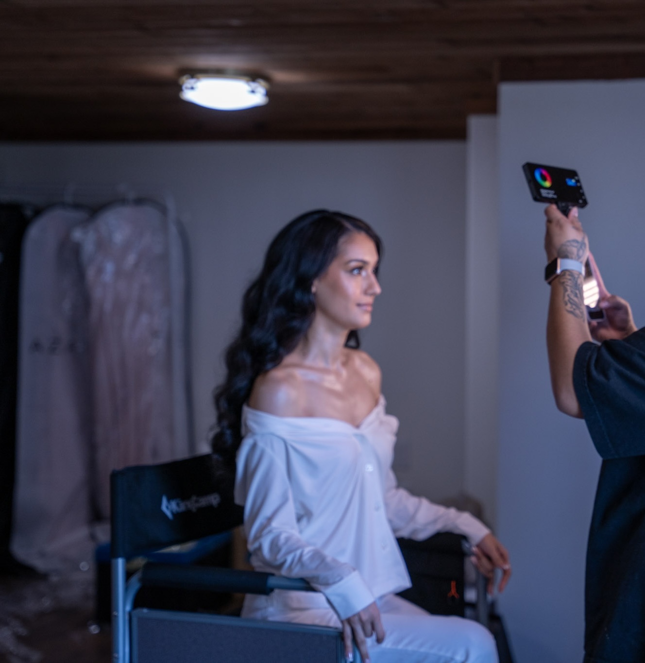 Woman with long dark hair taking a selfie indoors, wearing an off-shoulder white top, with a wedding dress in the background.