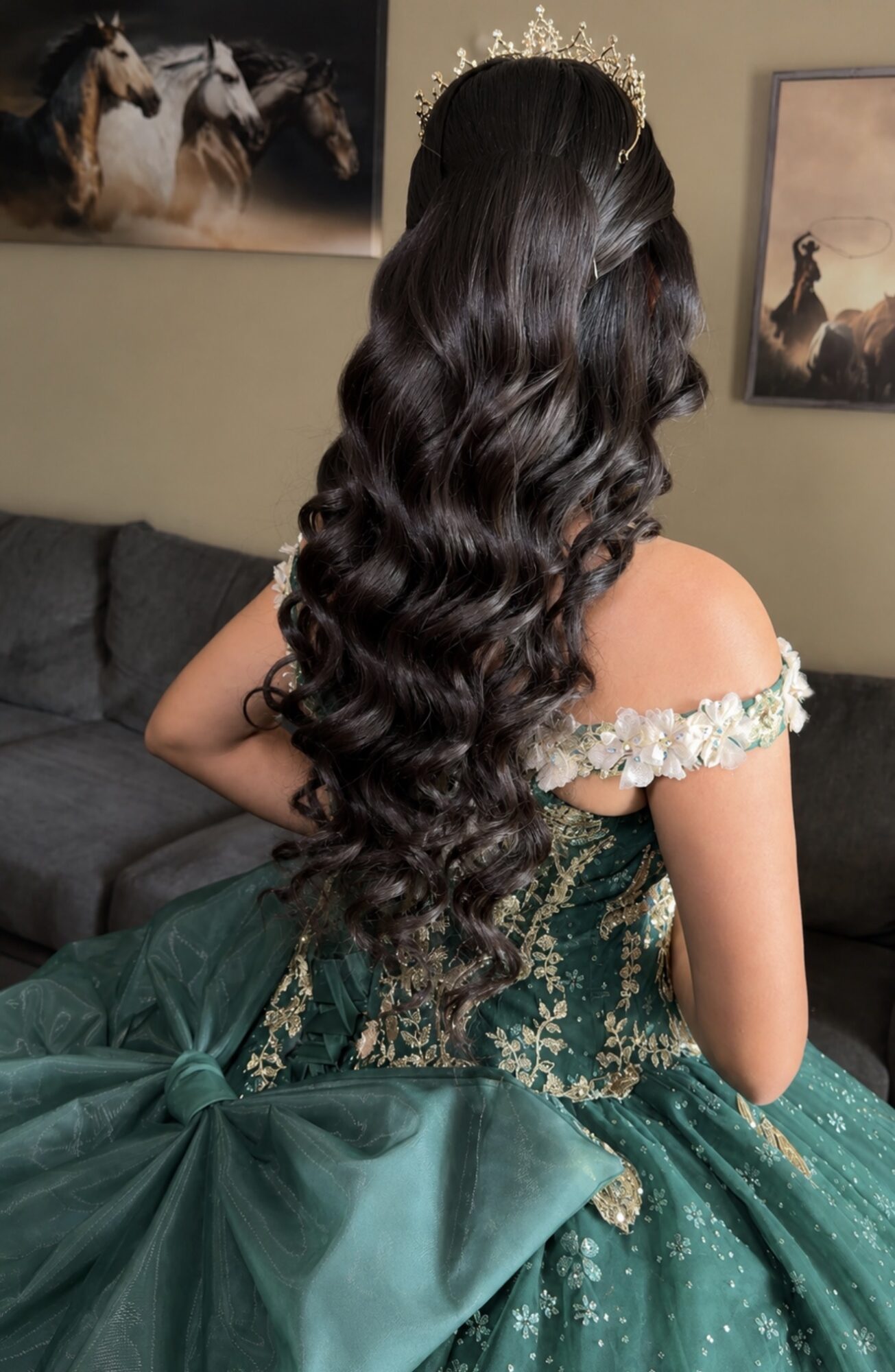 Woman with long, wavy dark hair wearing a green dress with floral details, sitting on a sofa in a room with horse-themed artwork.