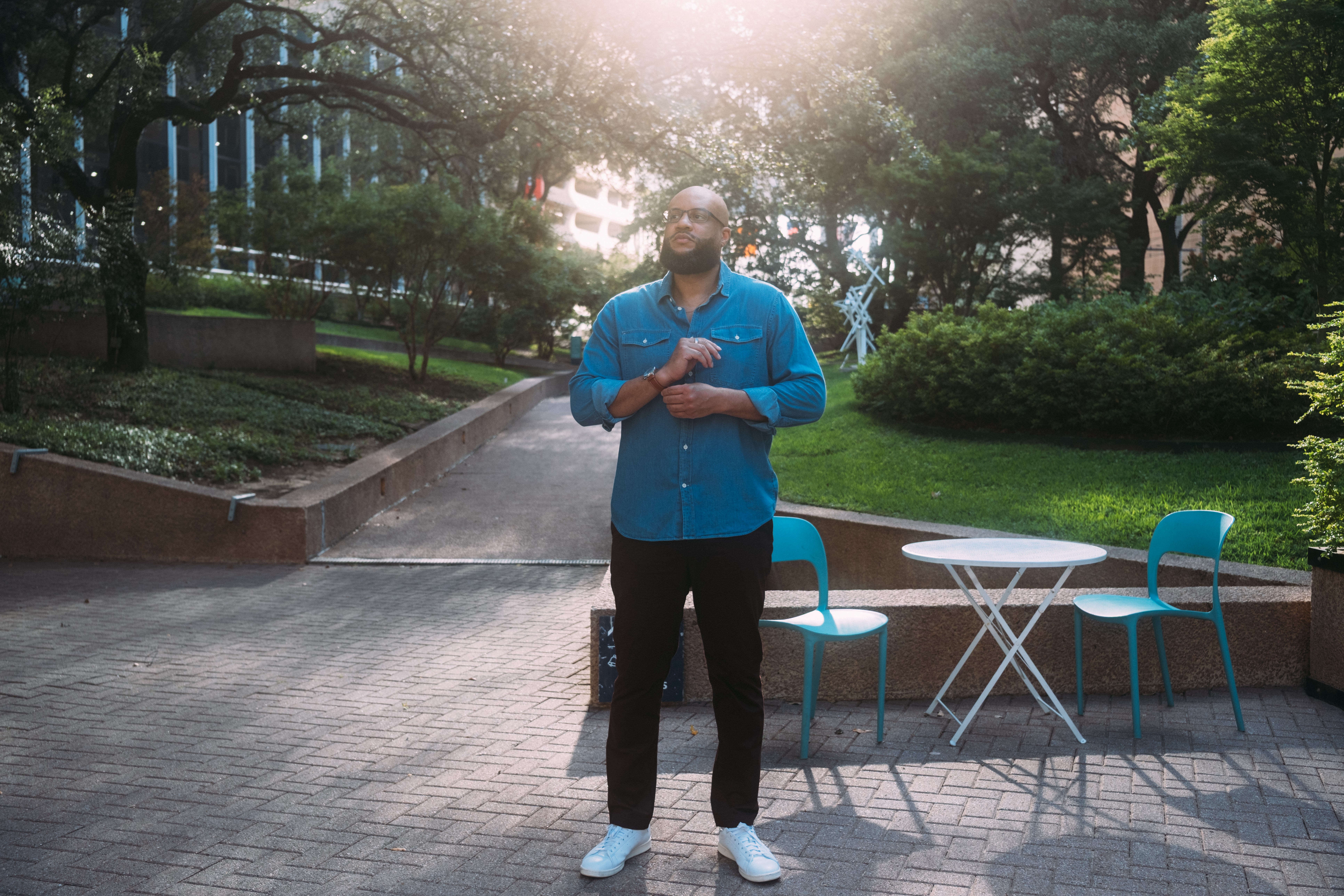Man standing outdoors near a table and chairs, with trees and sunlight in the background.