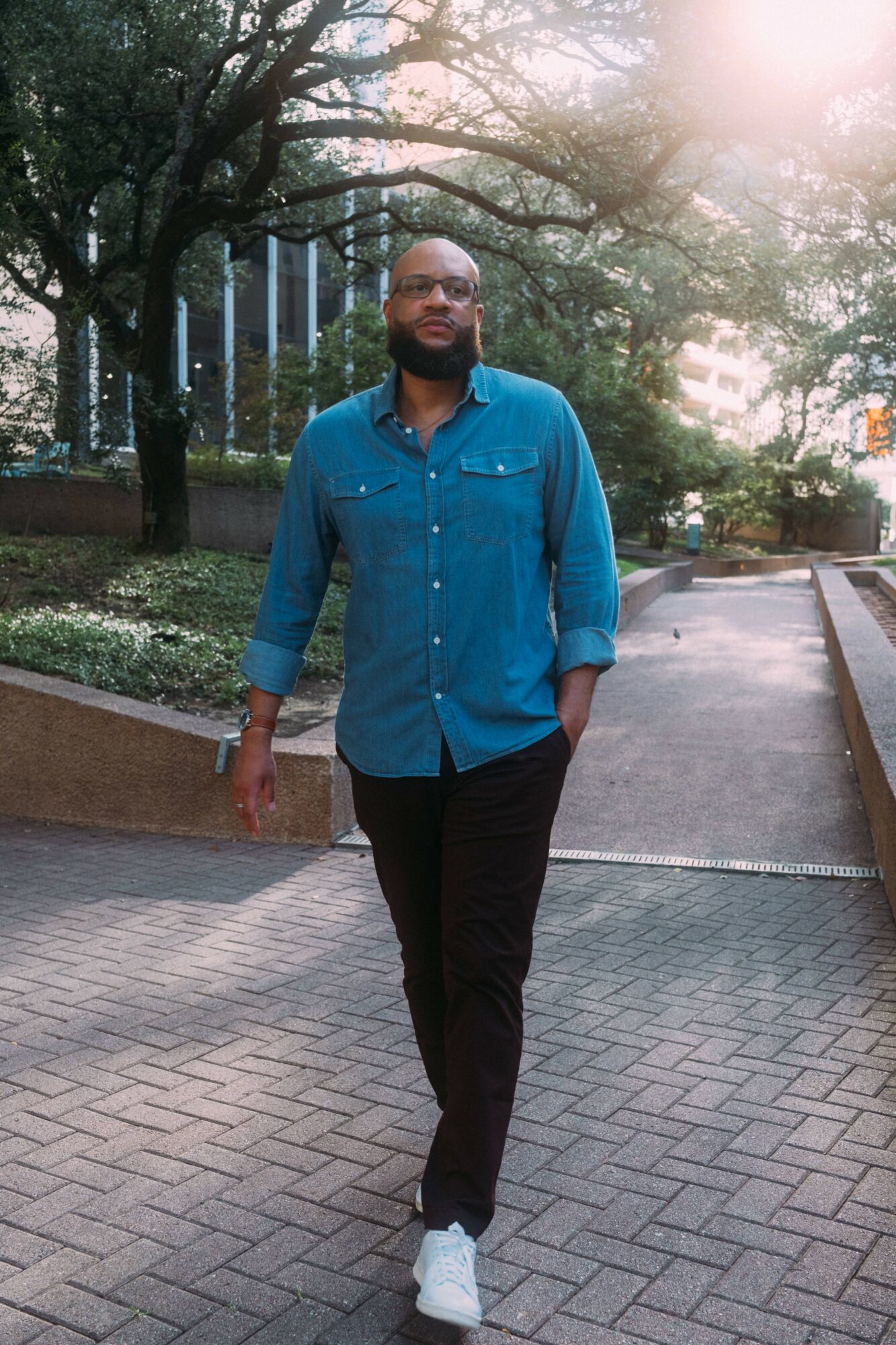 Man walking outdoors on paved path, trees and buildings in background, sunlight filtering through trees.