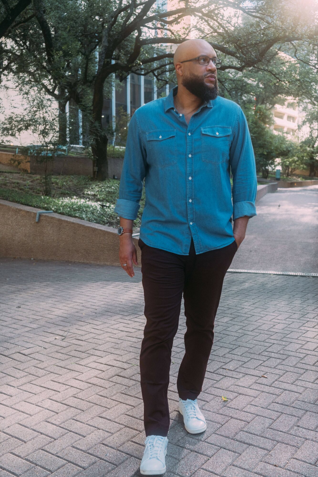 Man walking outdoors in a park with trees and buildings in the background, wearing a blue shirt, black pants, and white sneakers.