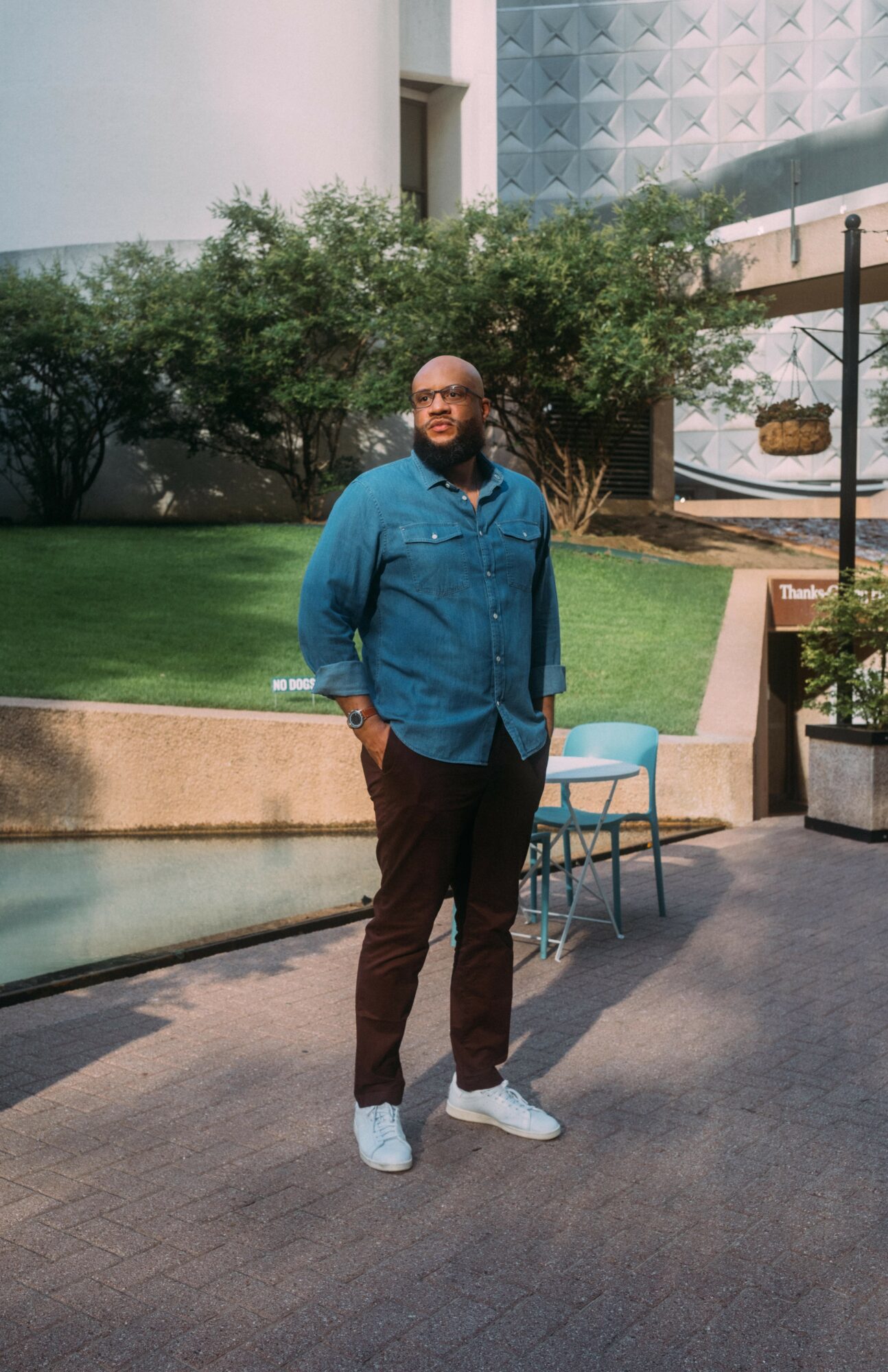 Man standing outdoors near a water feature, wearing a blue shirt, dark pants, and white sneakers, with greenery and modern buildings in background.