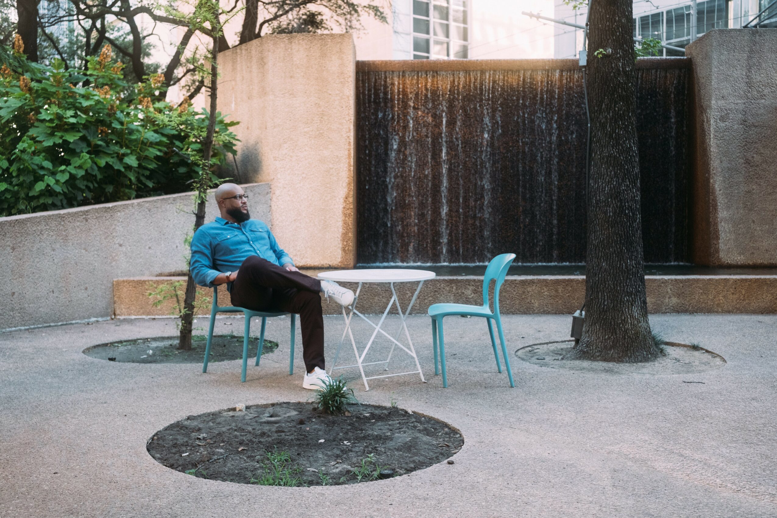 Person sitting at a table outdoors with two chairs, a tree, and a water feature in the background.