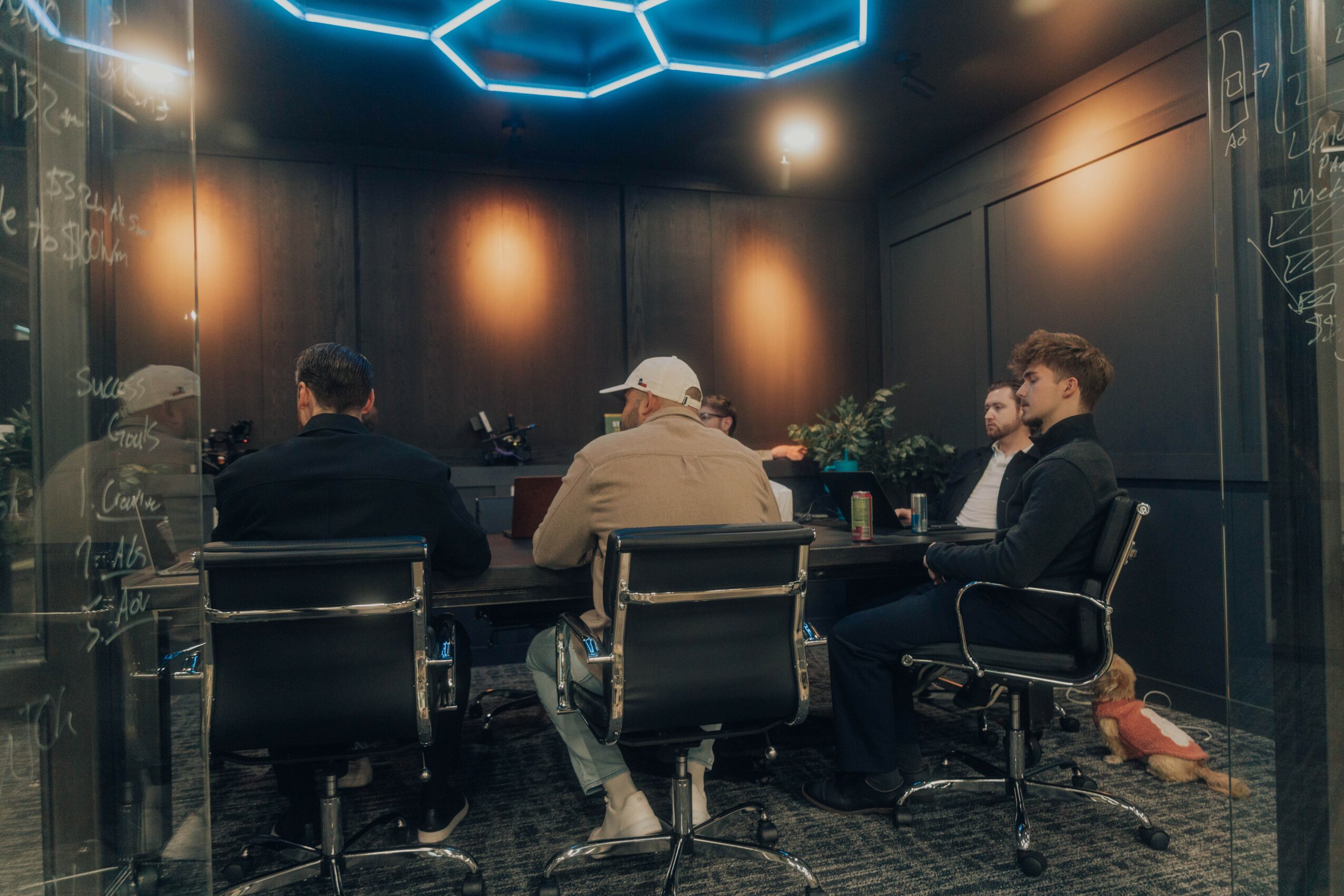 Four people seated around a table in a dark room with blue hexagonal ceiling lights, black walls, and a plant on the table.