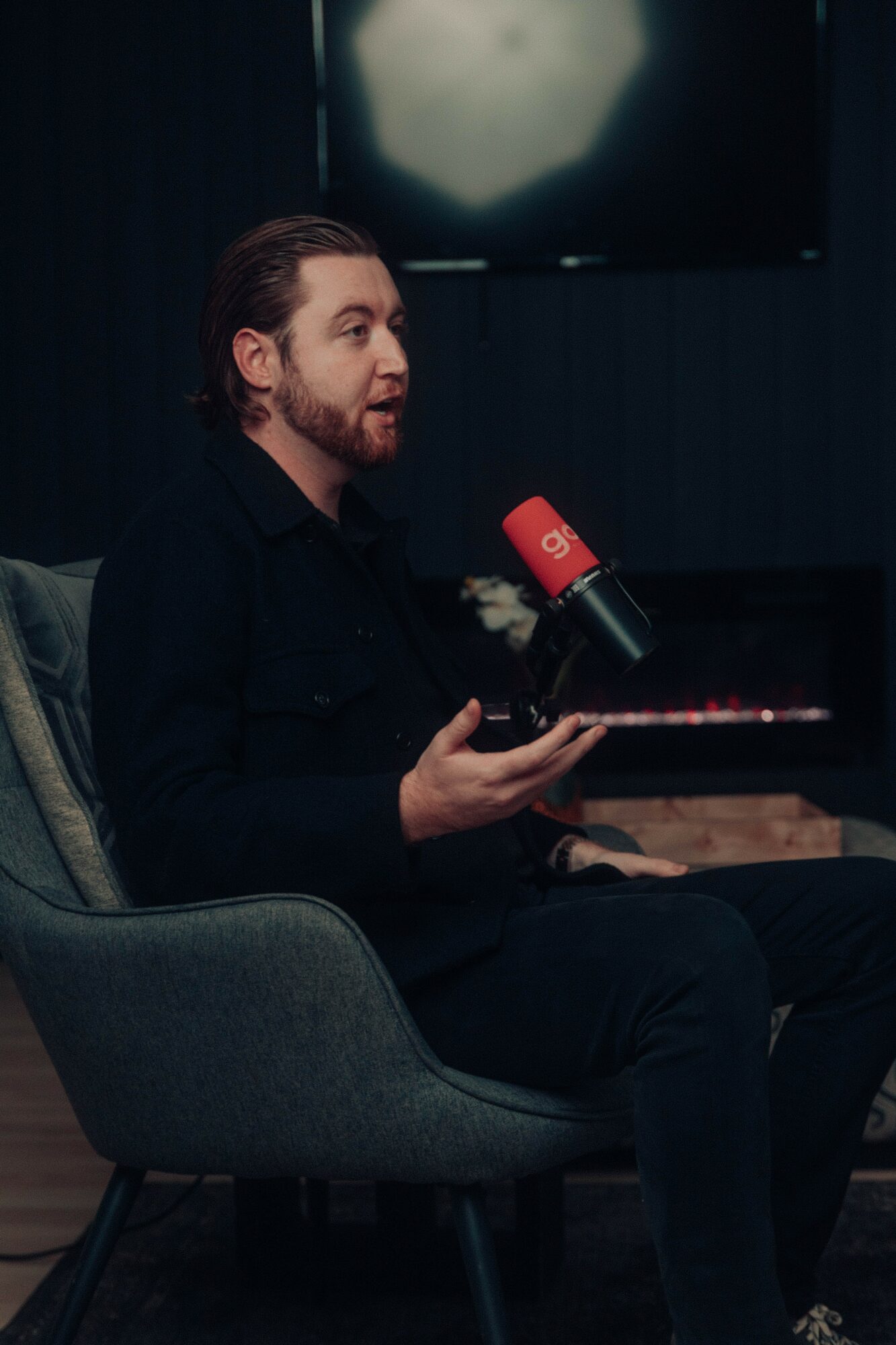 Man with beard and long hair speaking into a red microphone during an interview, sitting in a gray chair.
