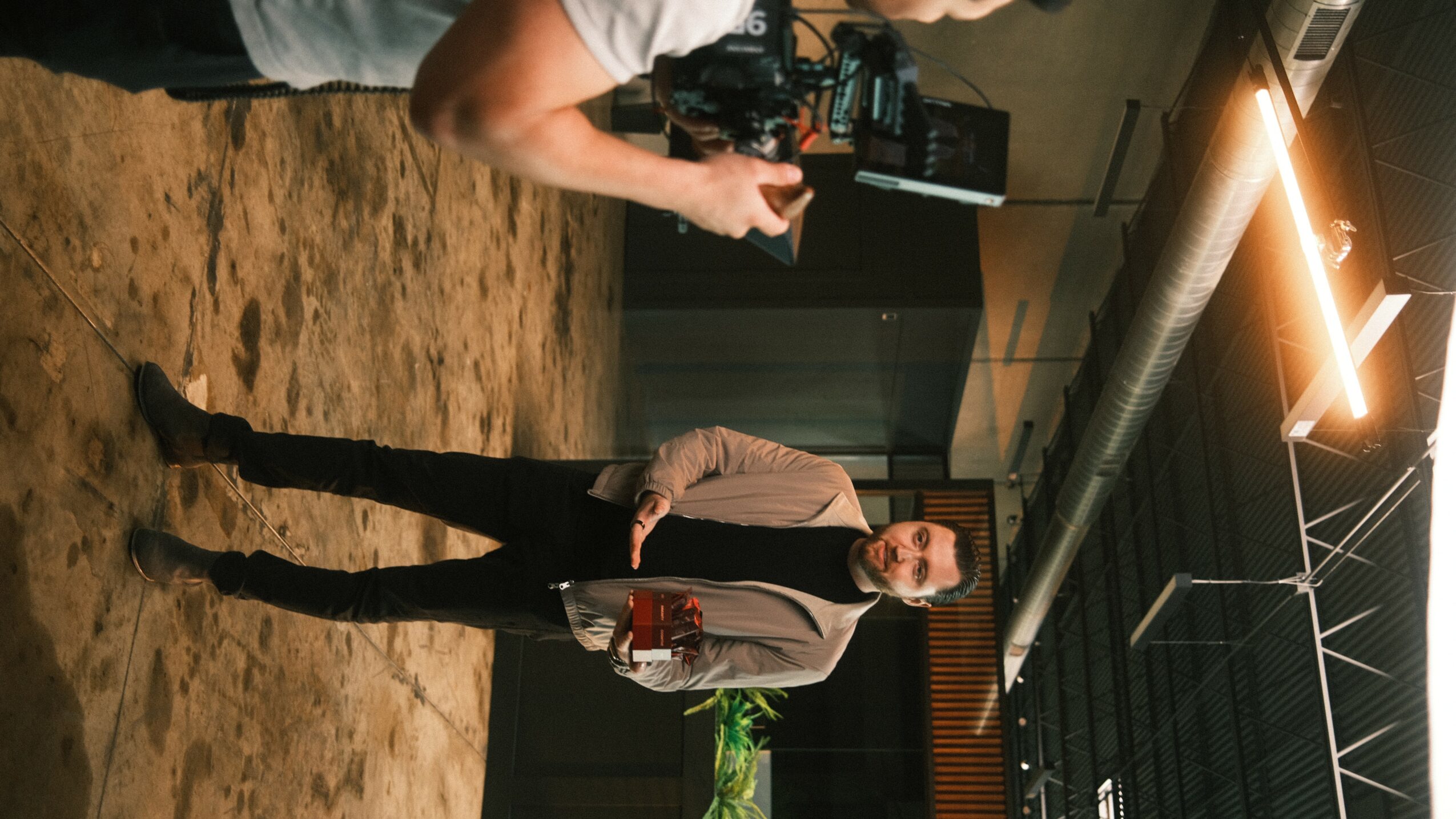 Man in a suit standing in a room with a camera operator filming him, with industrial ceiling and lighting.