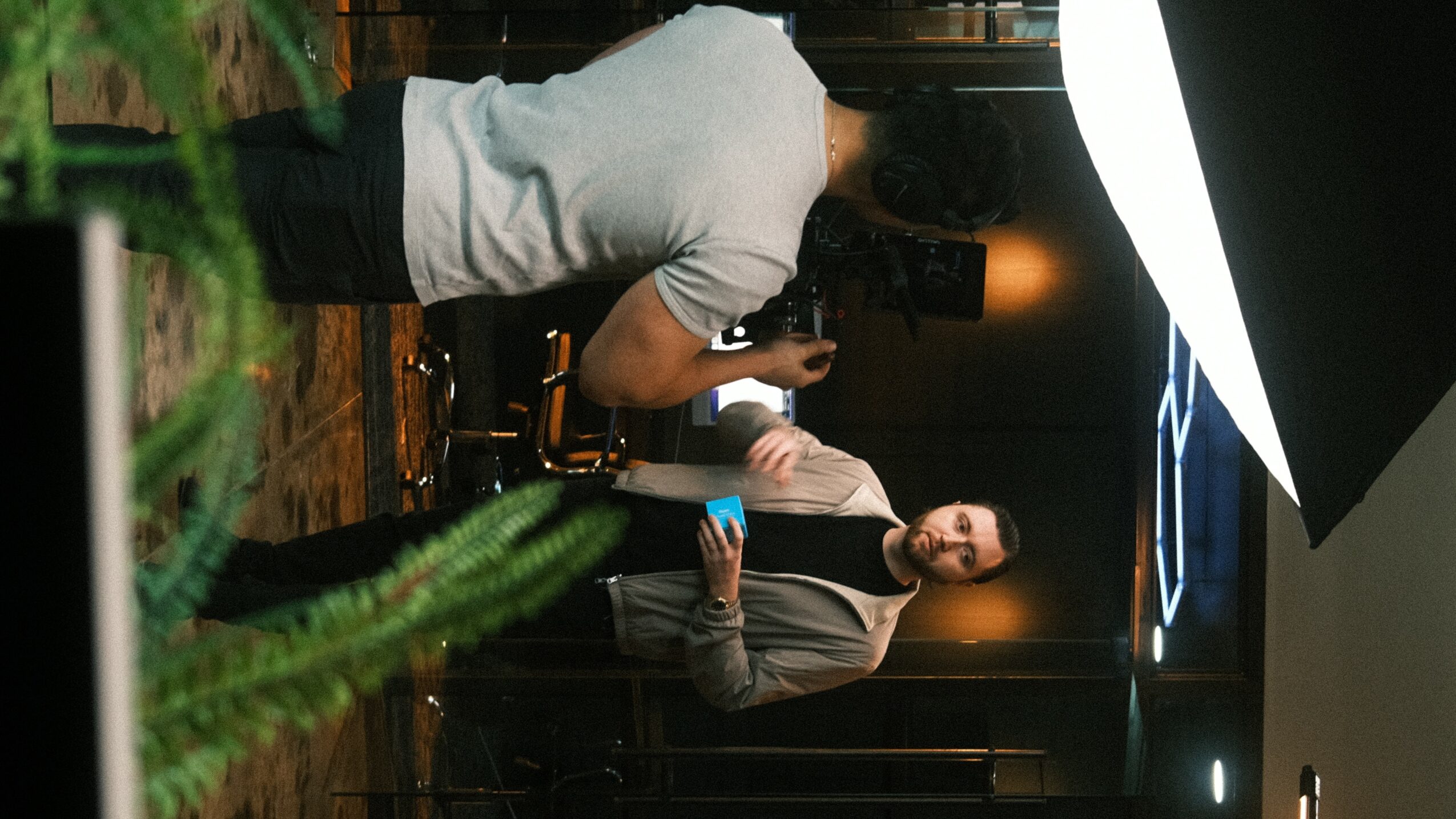 Two people in a studio or interview setting, one holding a microphone and the other sitting, with studio lighting and equipment visible.