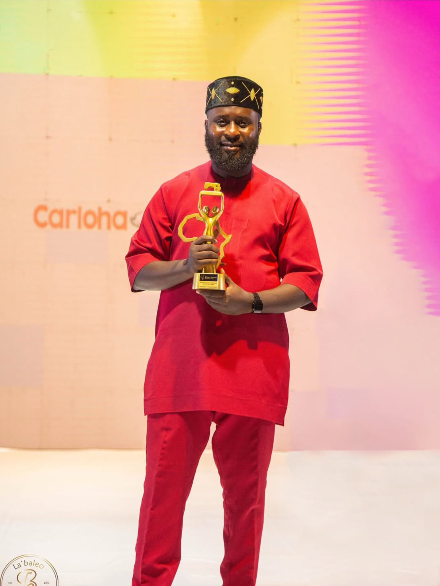 Man in red traditional attire holding a trophy, smiling, standing on stage with colorful background.