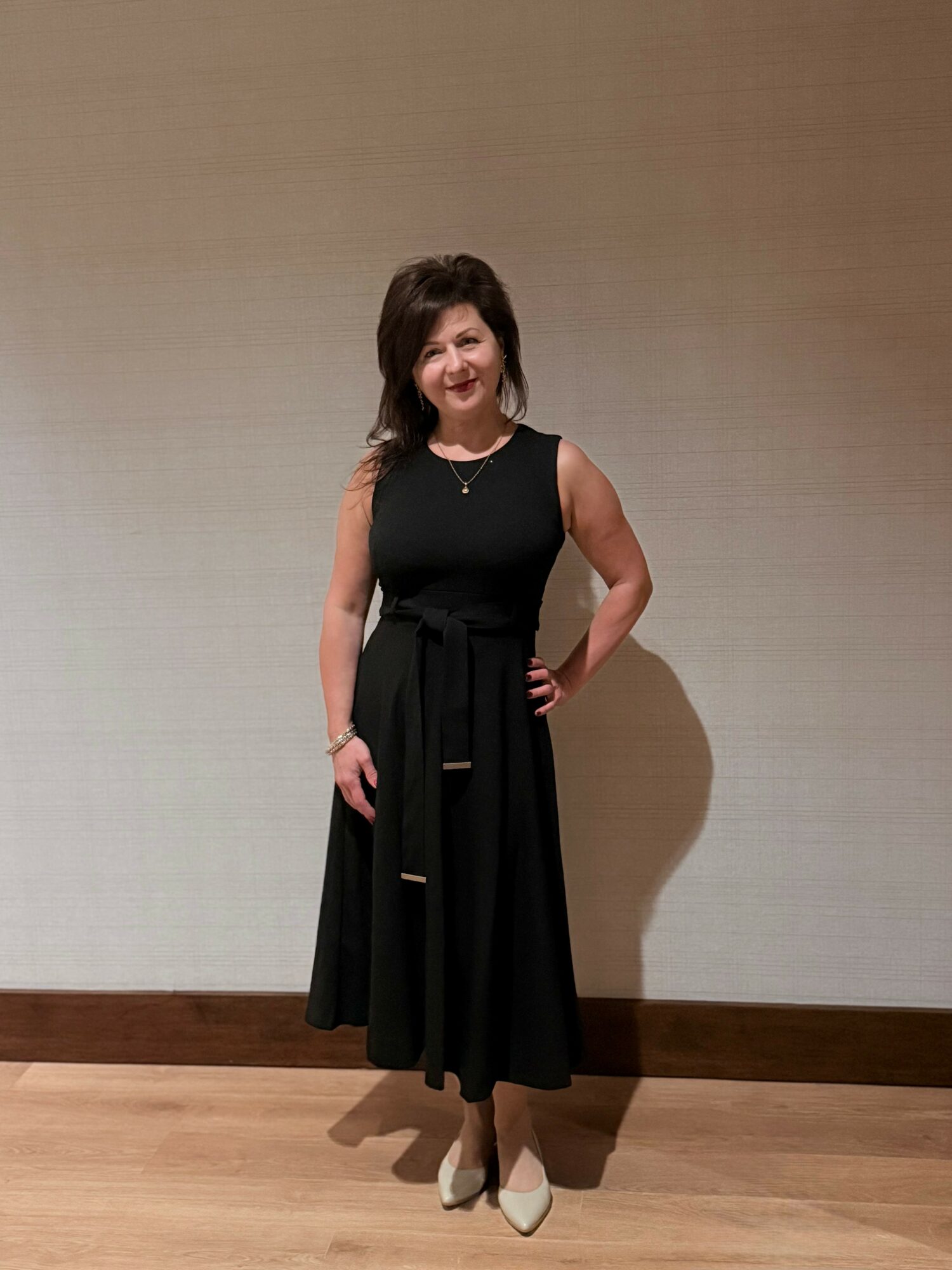 Woman in a black dress standing indoors, smiling, with one hand on her hip, against a plain wall.