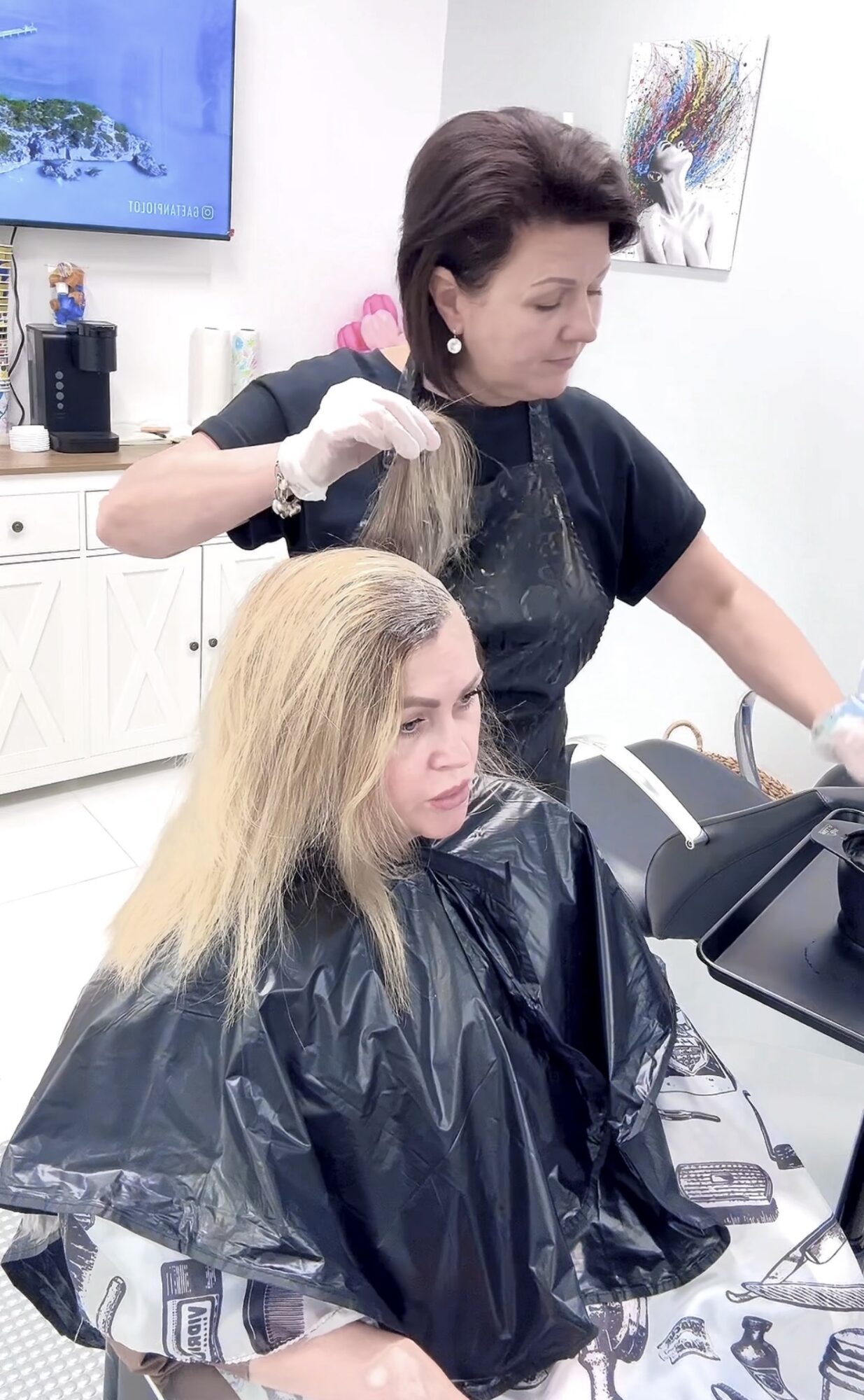 Woman applying hair dye to a seated woman with blonde hair in a salon, both wearing black capes.