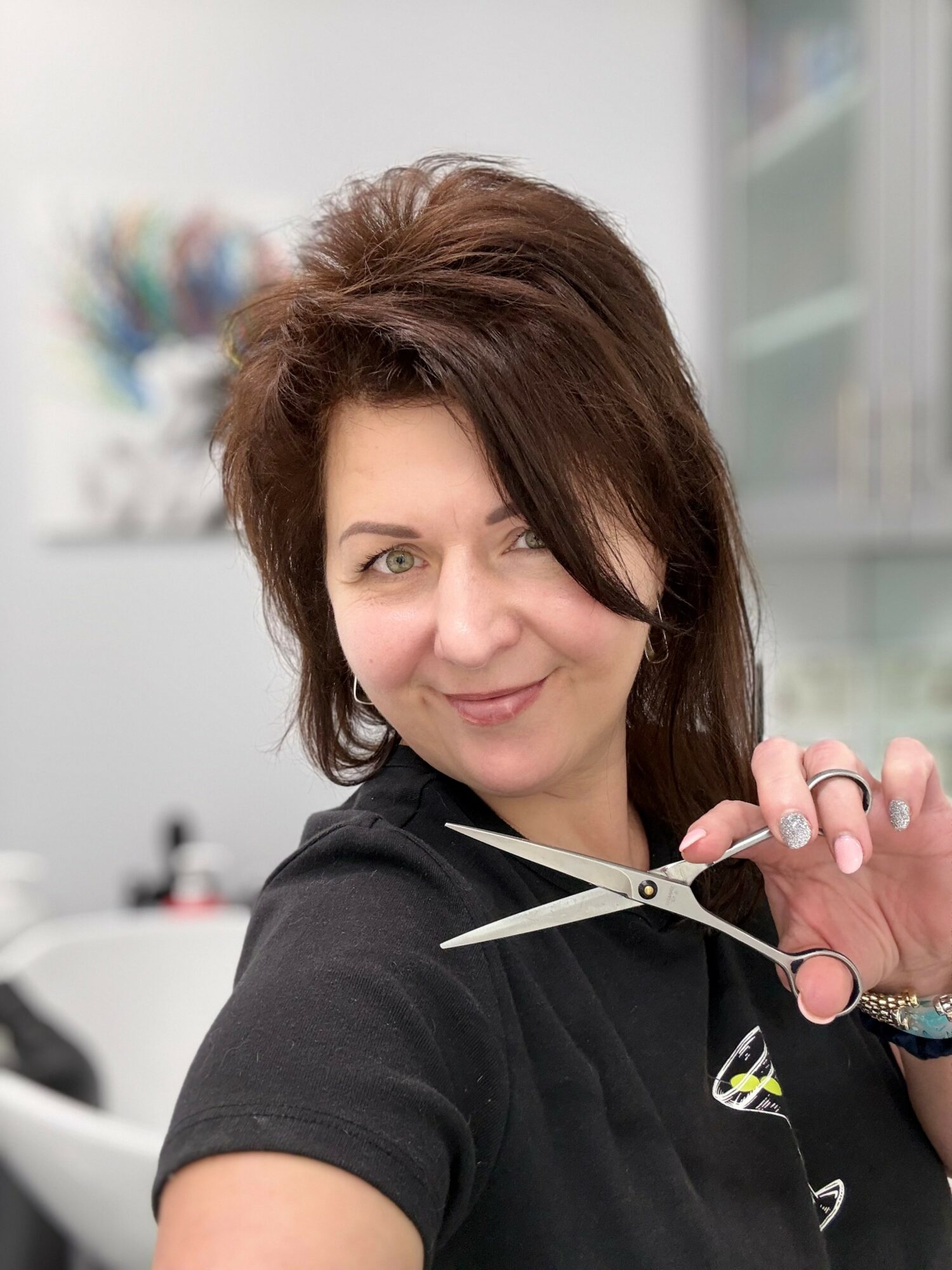 Woman with brown hair smiling, holding scissors, in a bright indoor setting.
