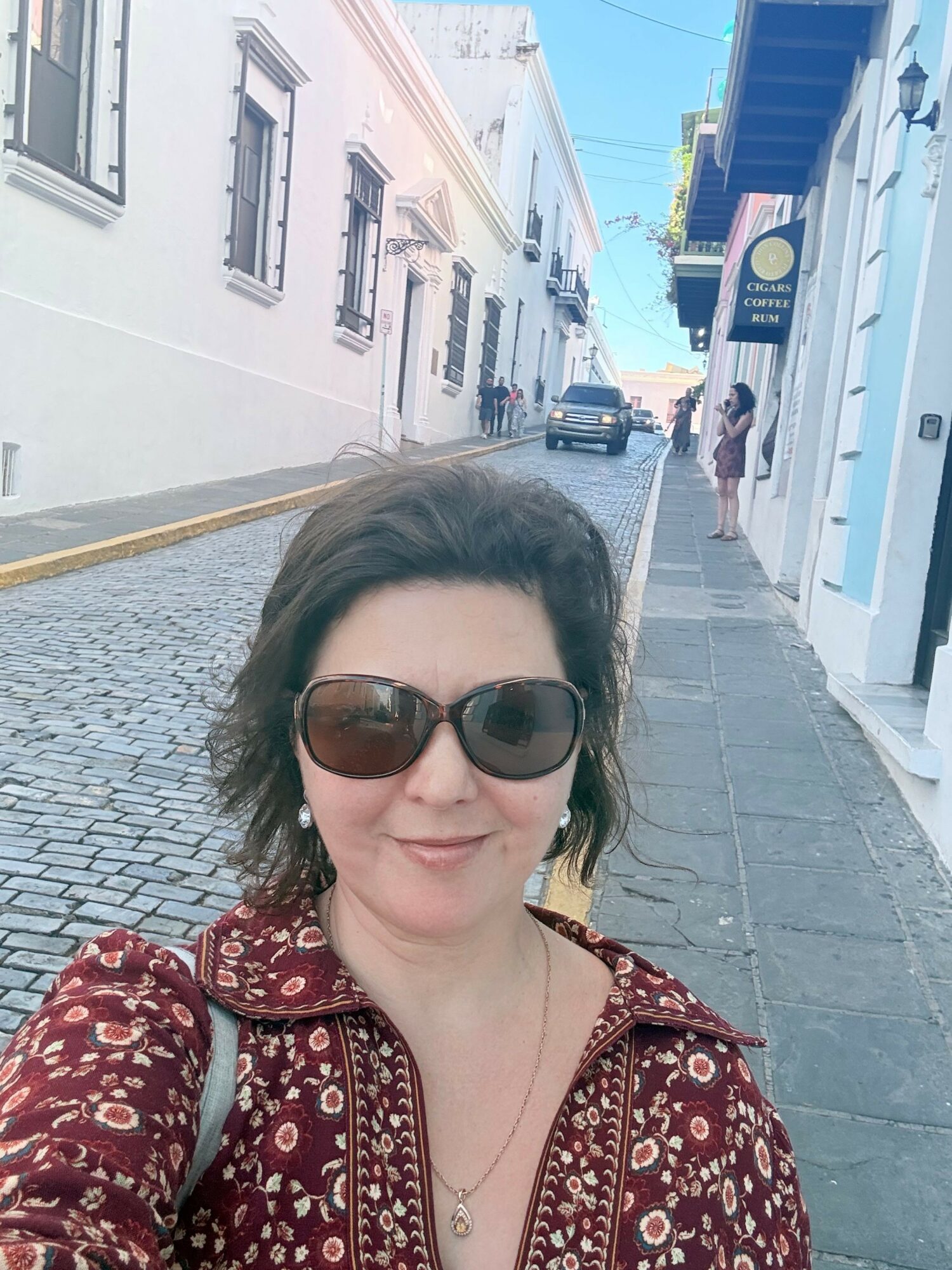 Woman with sunglasses and patterned top taking a selfie on a cobblestone street, buildings and a blue sky in background.