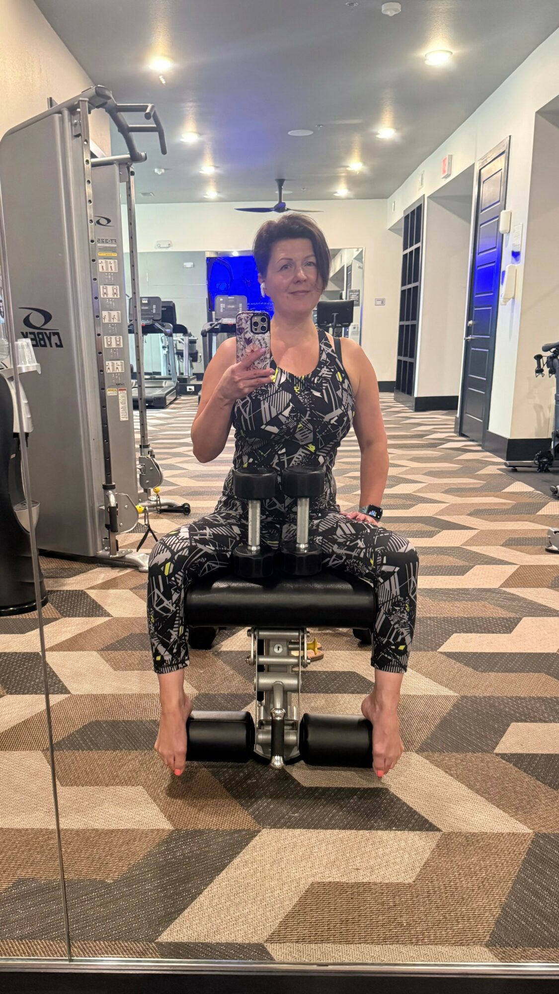 Woman taking a selfie in a gym mirror, sitting on a workout machine, wearing patterned workout clothes, with gym equipment in background.