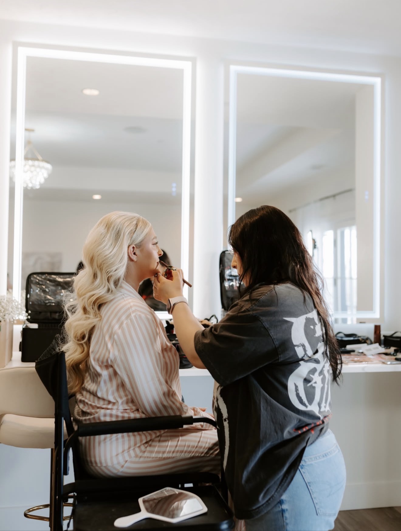 Two women in a makeup room, one applying makeup to the other, with large mirrors and bright lighting behind them.
