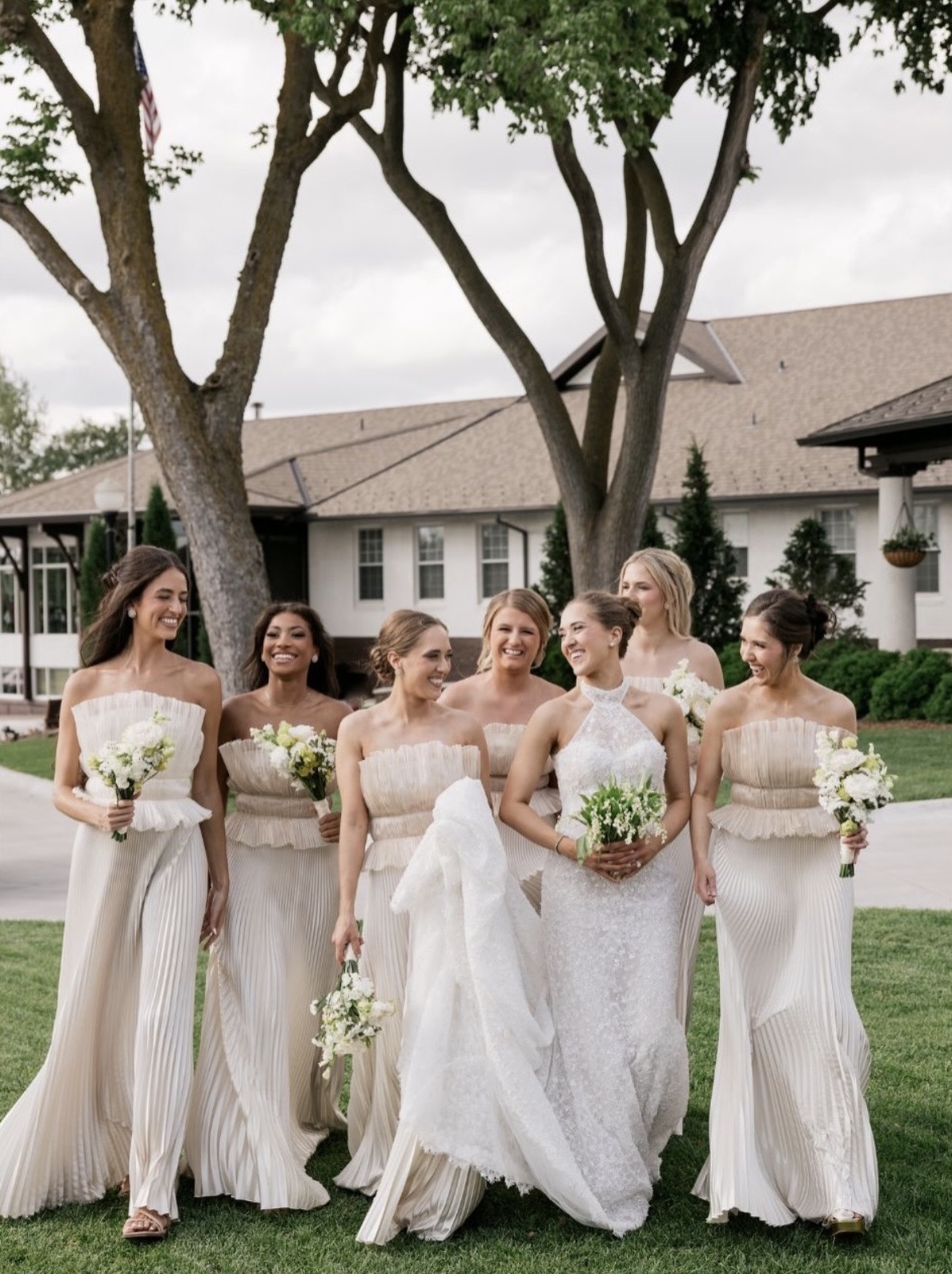 Six women in white dresses holding bouquets, standing outdoors in front of trees and a building, smiling.