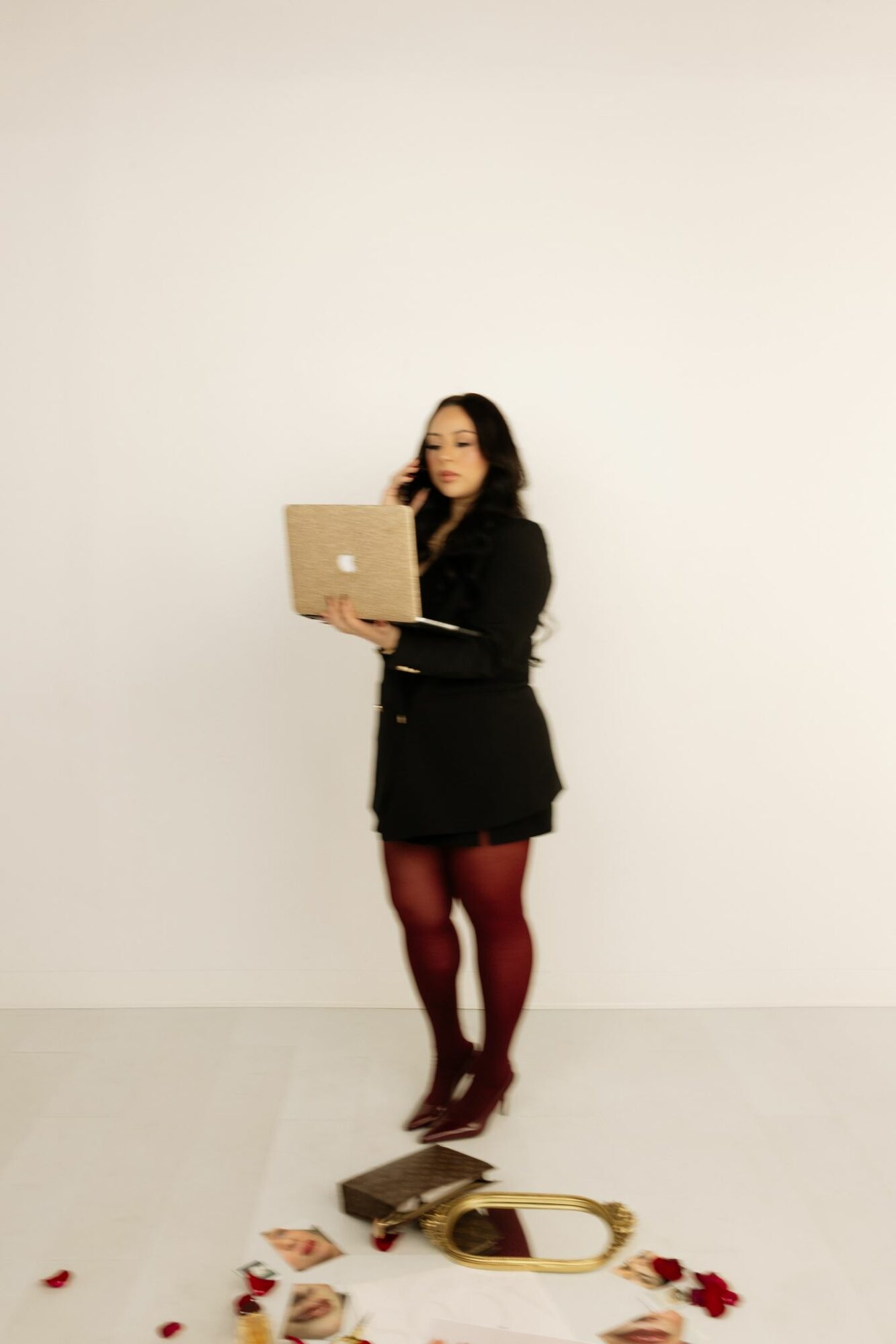 Woman standing against a plain white wall, holding a laptop, with a mirror and rose petals on the floor.