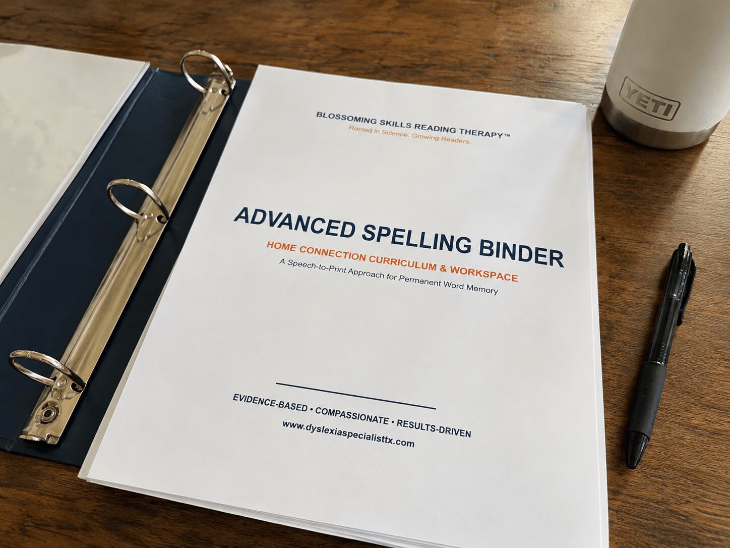 Open binder with a printed document titled 'Advanced Spelling Binder' on a wooden table, with a pen and a cup nearby.
