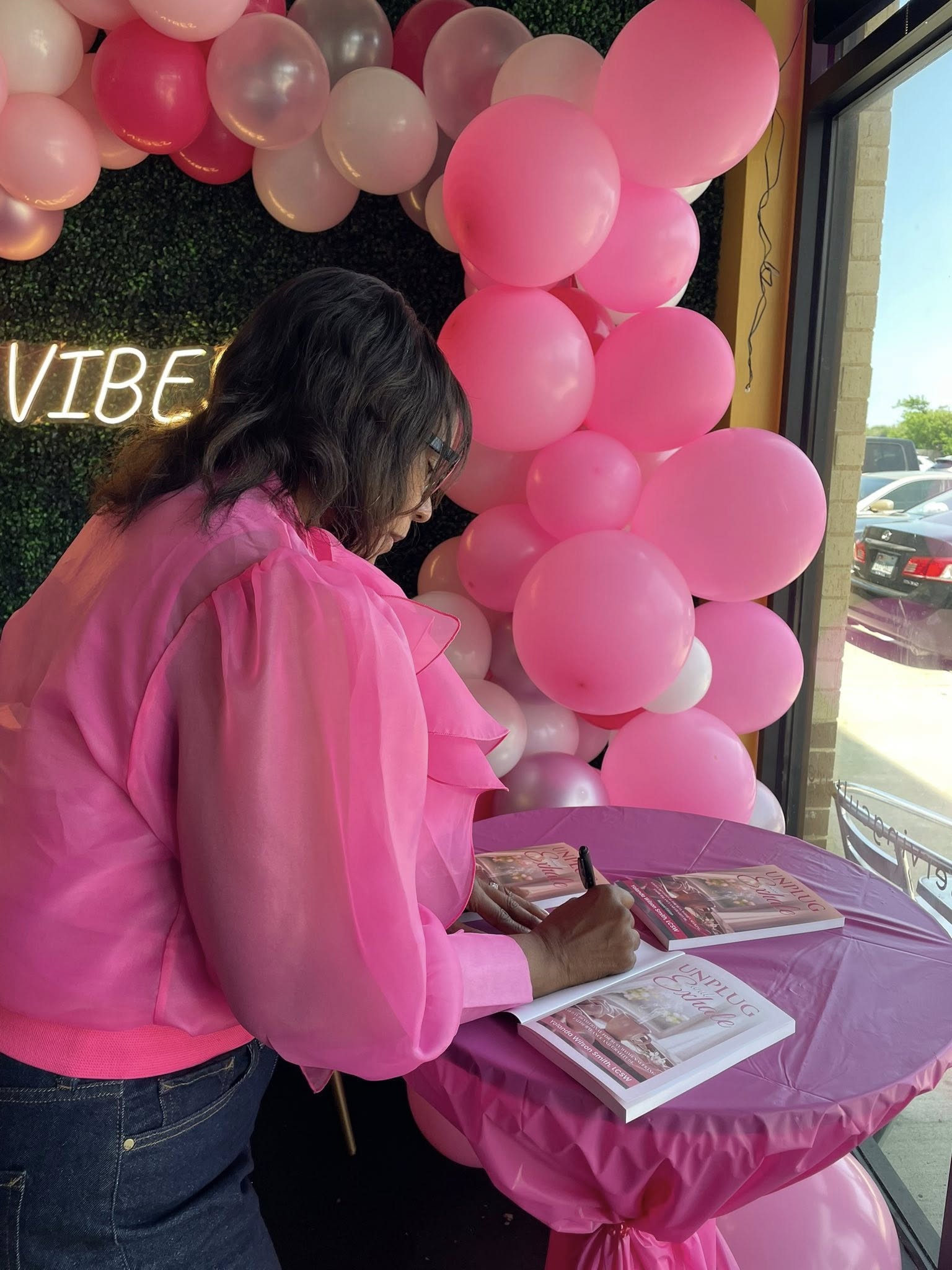 Woman in pink blouse writing in a notebook at a pink table with balloons outside window.