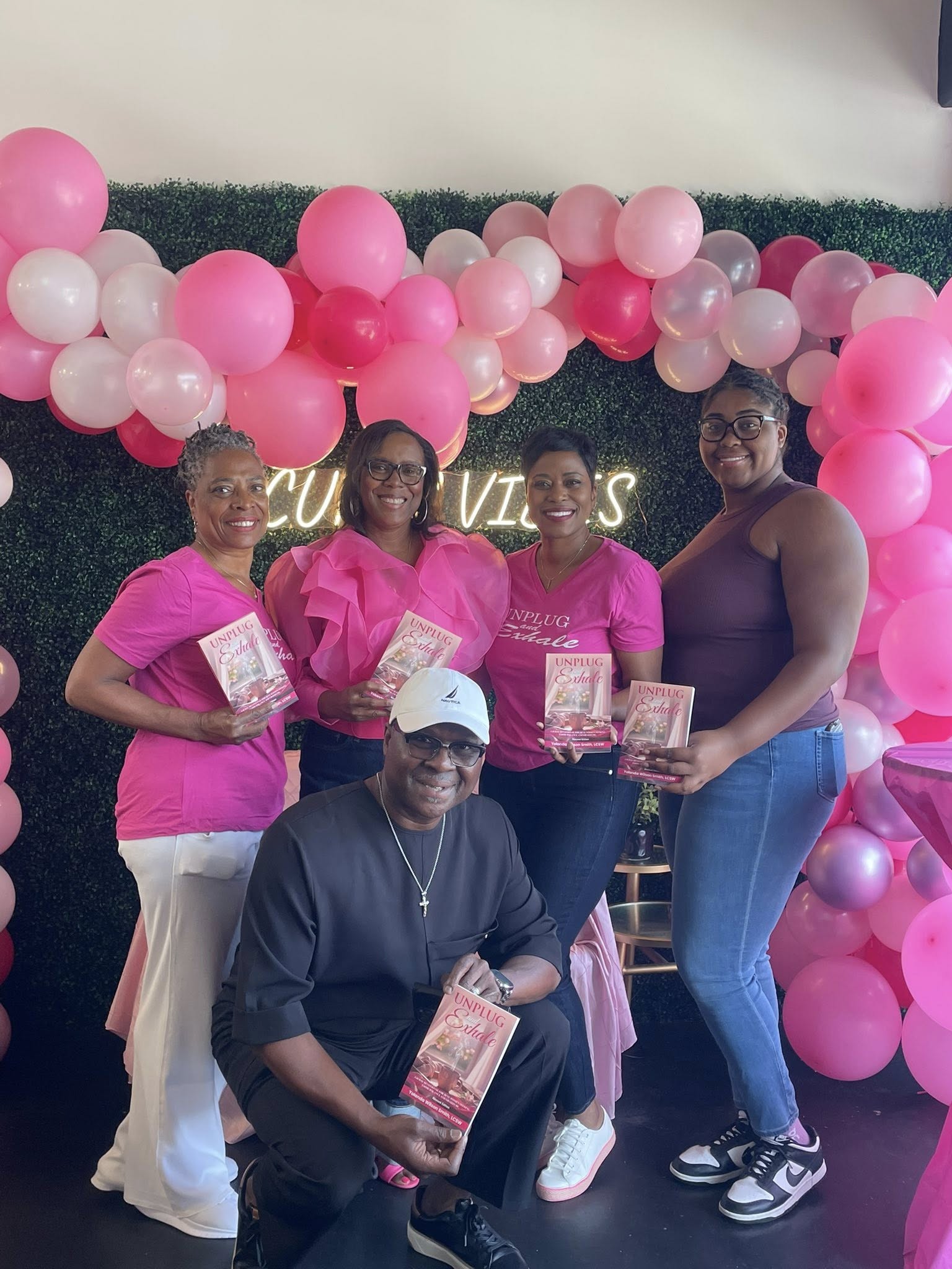 Four women and one man pose in front of pink and white balloons, holding books or pamphlets, at a celebration.
