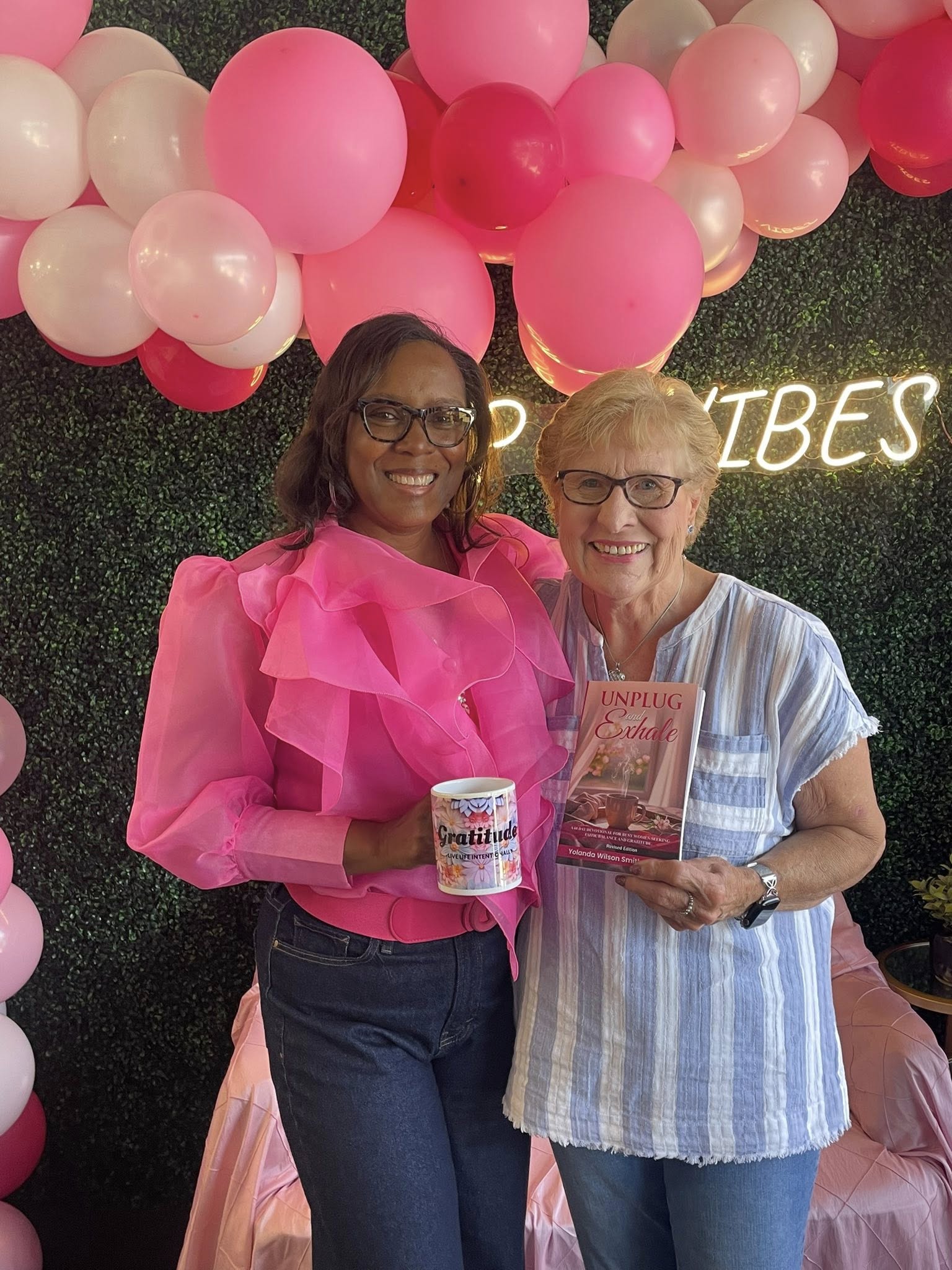 Two women smiling, one holding a book and a cup, standing in front of pink and white balloons and a green backdrop.