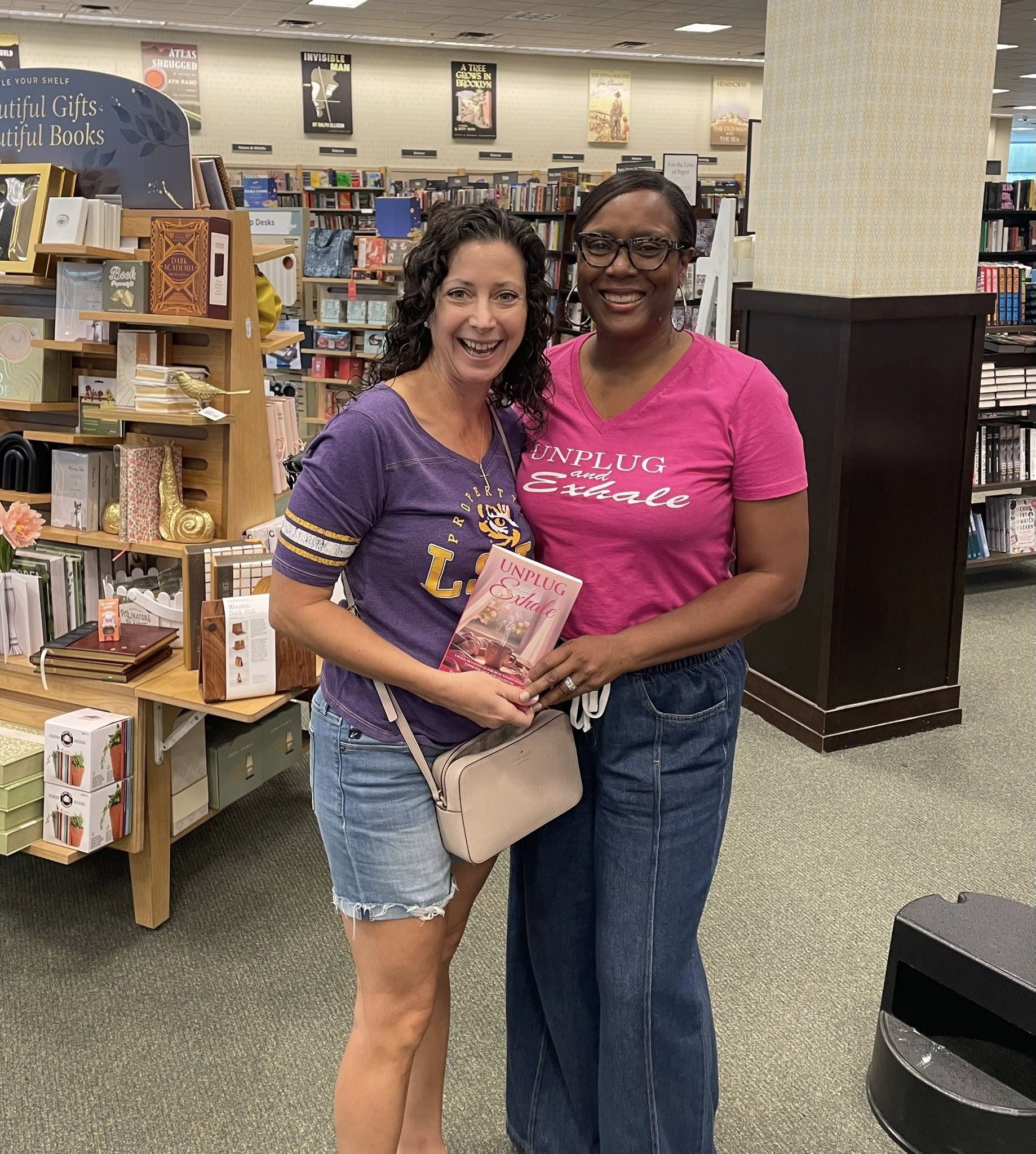 Two women smiling and holding a book in a bookstore, surrounded by shelves of books.
