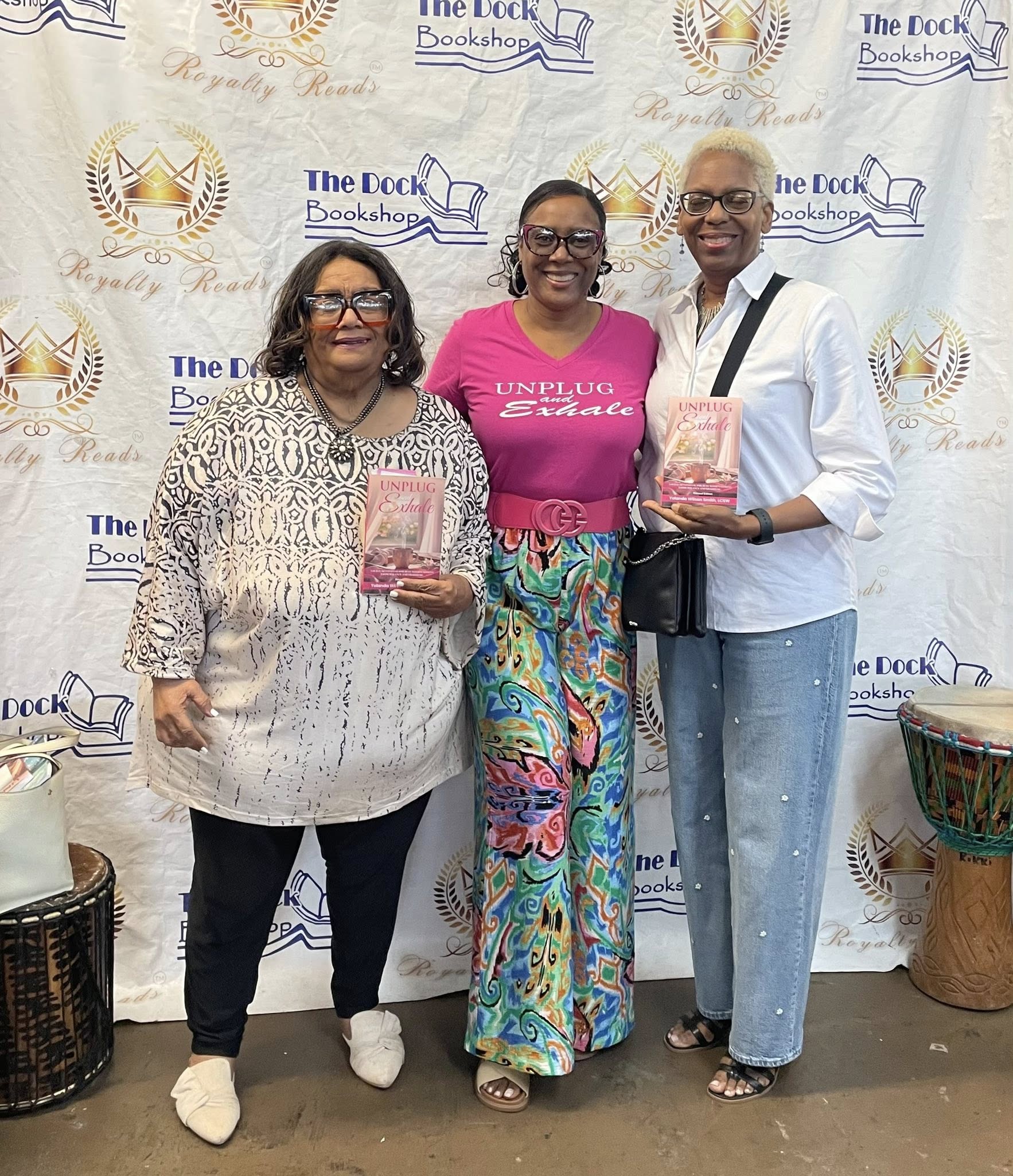 Three women standing together in front of a backdrop, holding books, smiling, with drums on the floor.