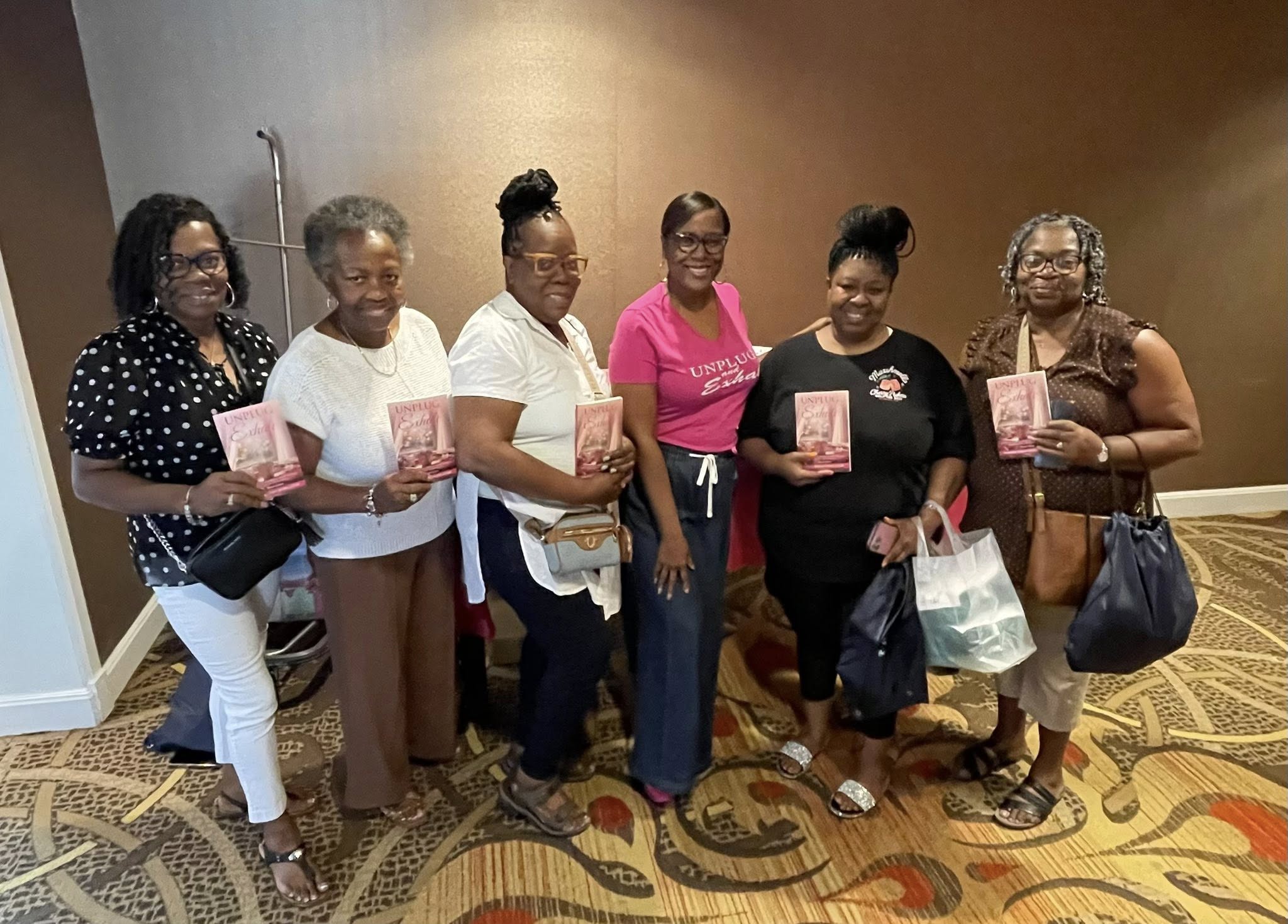 Group of seven women standing indoors, holding pink booklets and bags, smiling for the photo.
