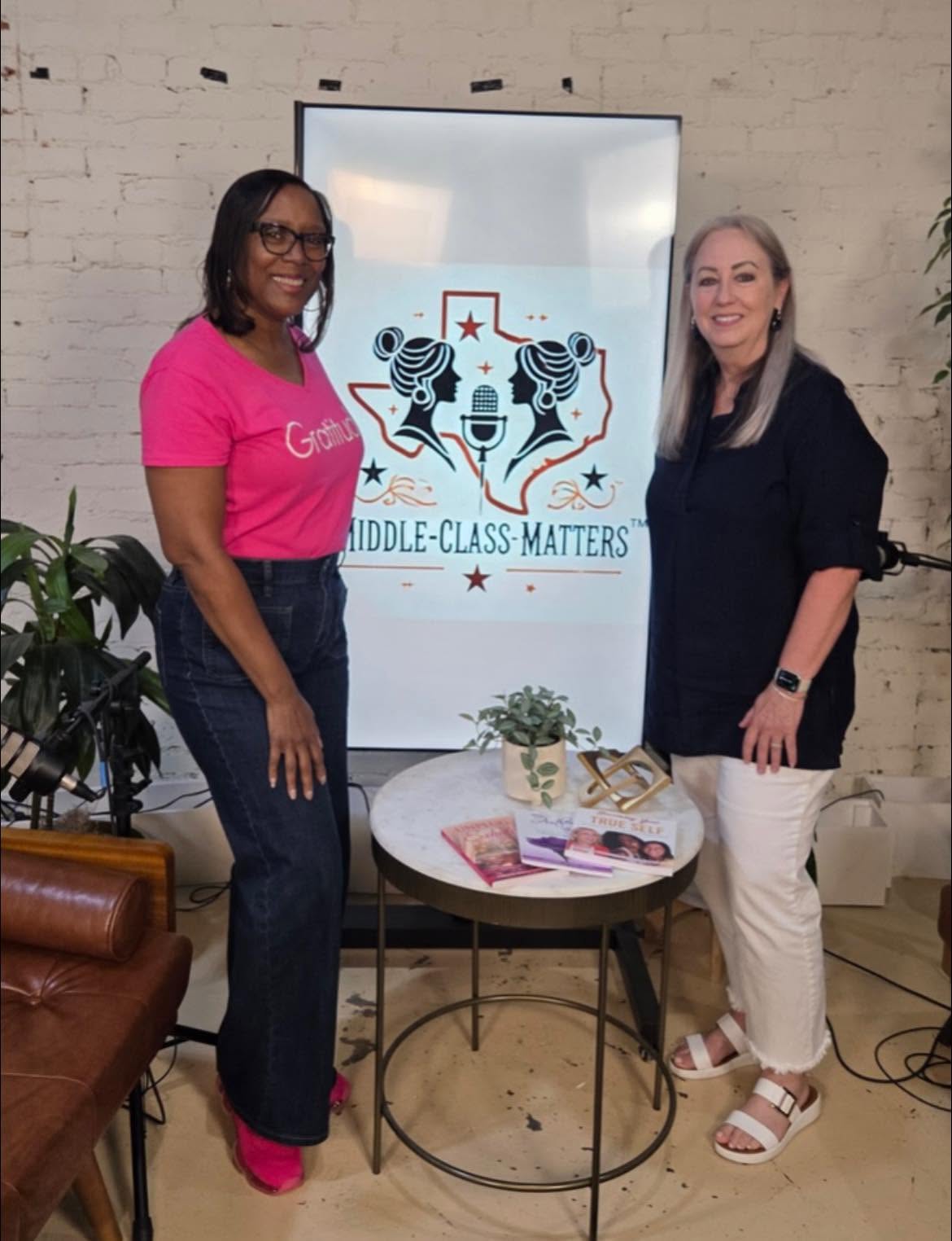 Two women stand beside a table with a plant and magazines, smiling in front of a sign that reads 'Middle-Class Matters'.
