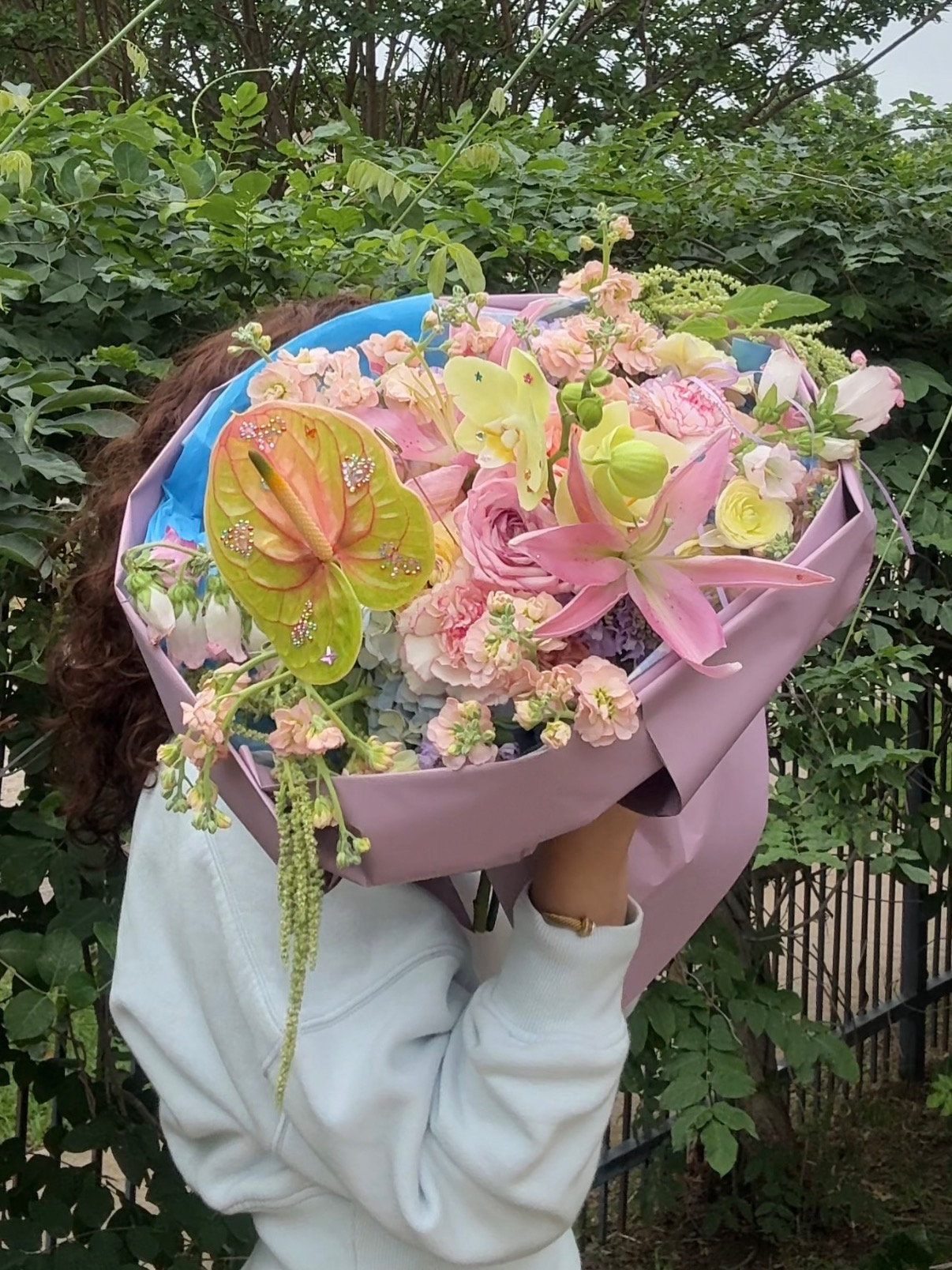Person holding a large bouquet of pink, yellow, and white flowers outdoors, with greenery in the background.