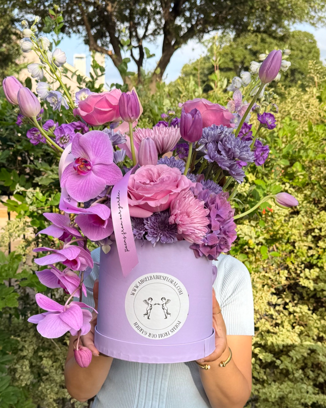 Person holding a purple hat filled with pink and purple flowers outdoors, trees and greenery in background.