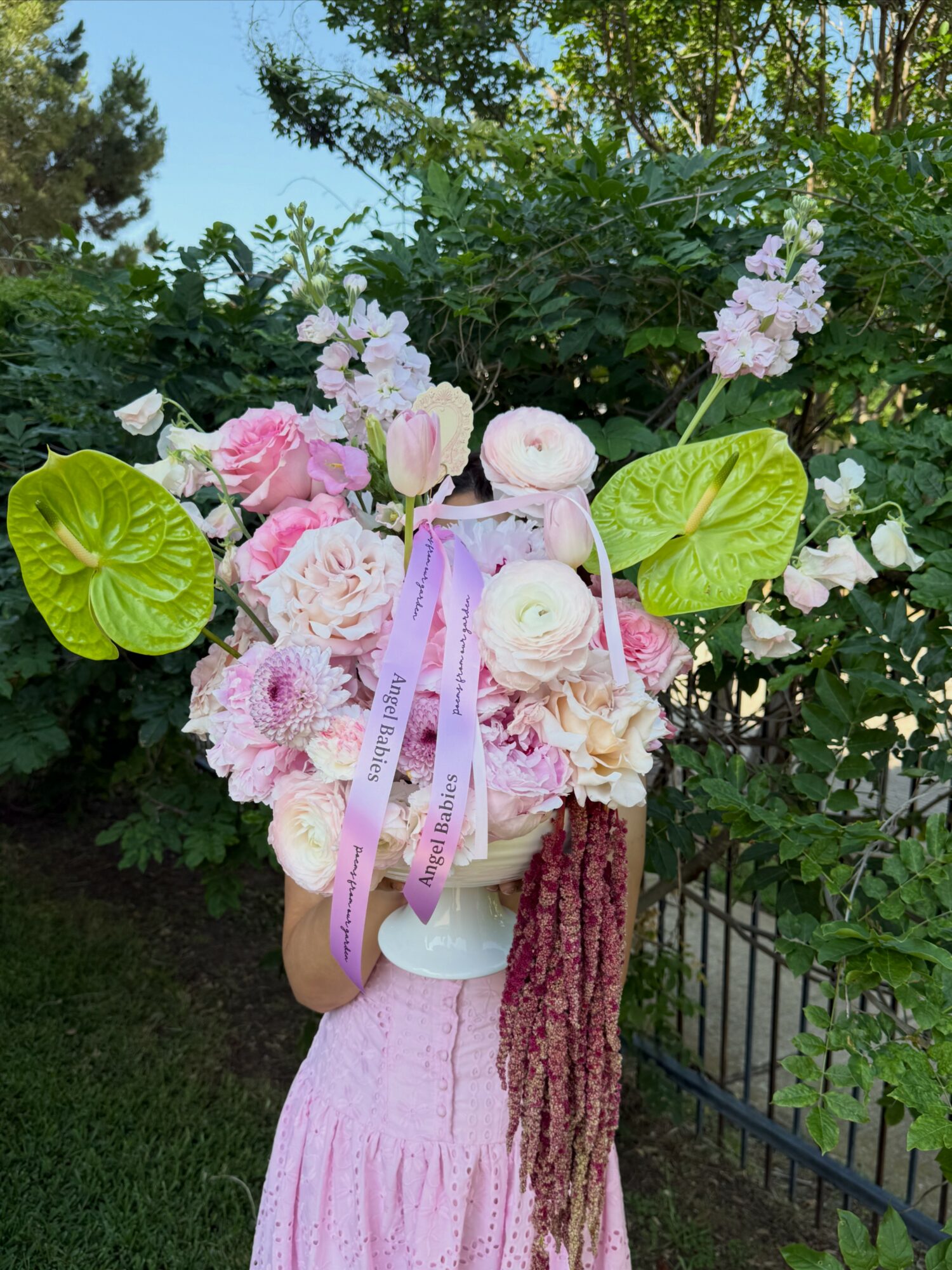 Person holding a pink and white flower bouquet with green leaves, outdoors with trees and a fence in background.