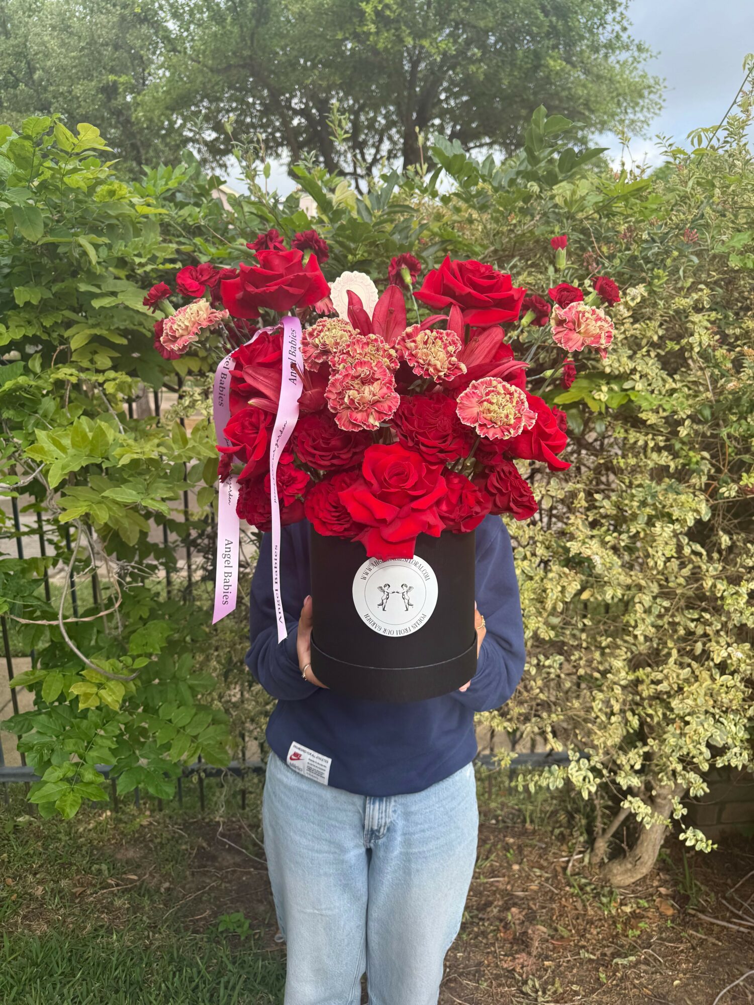 Person holding a large bouquet of red and pink flowers outdoors, with trees and greenery in the background.