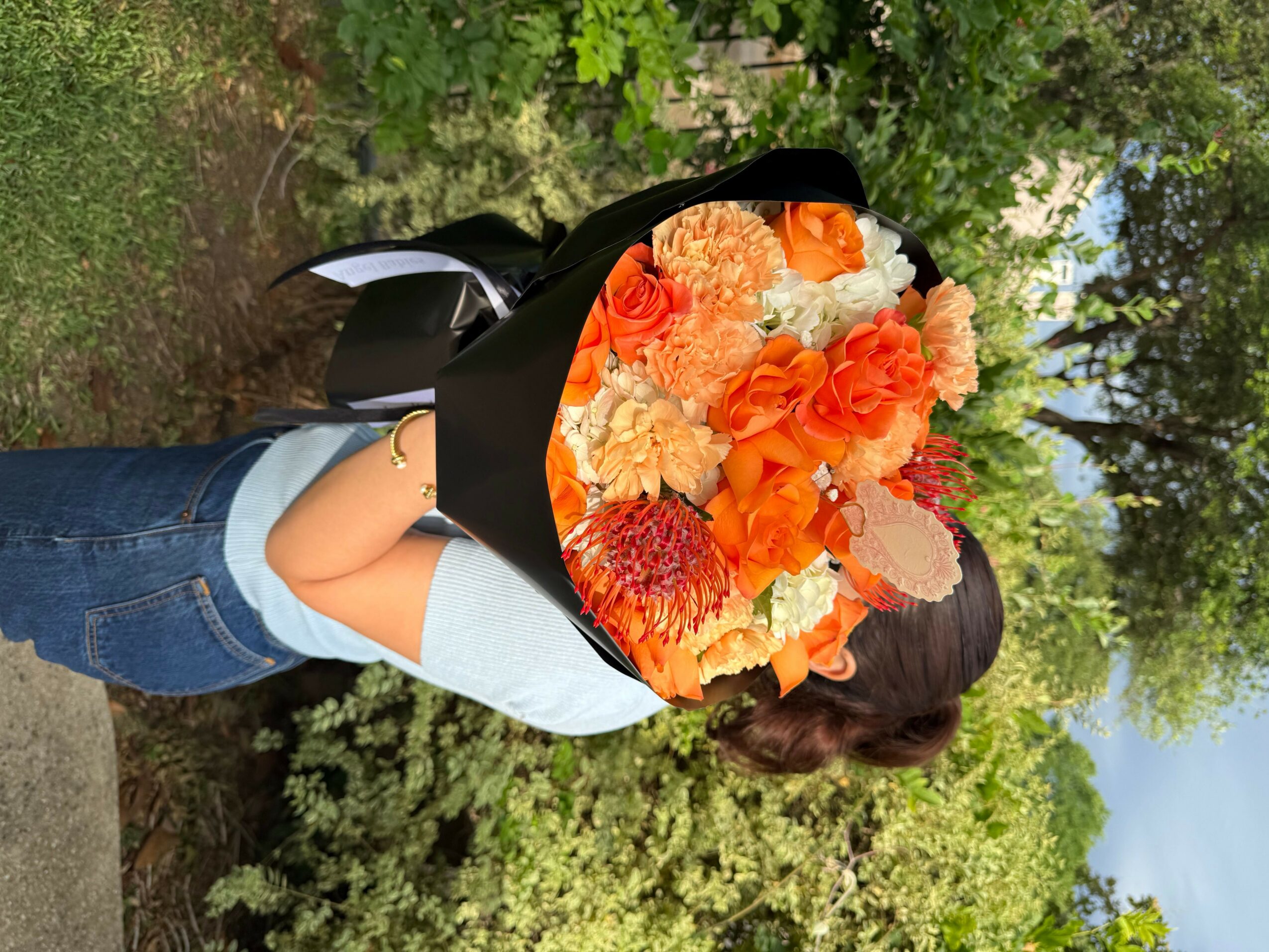 Person holding a large bouquet of orange, white, and red flowers outdoors with trees and greenery in the background.