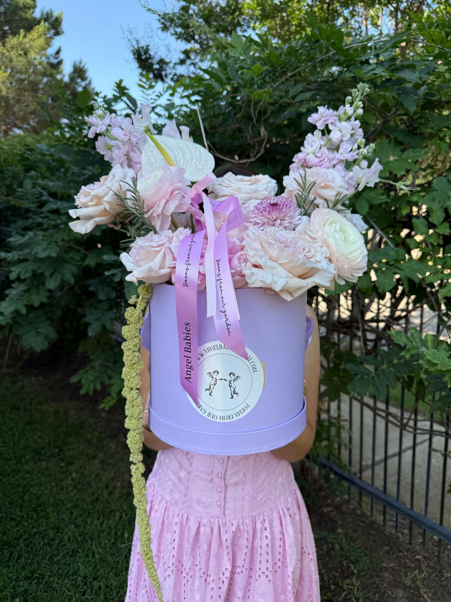 Person holding a lavender gift box with a pink ribbon and flowers, standing outdoors near a fence and greenery.