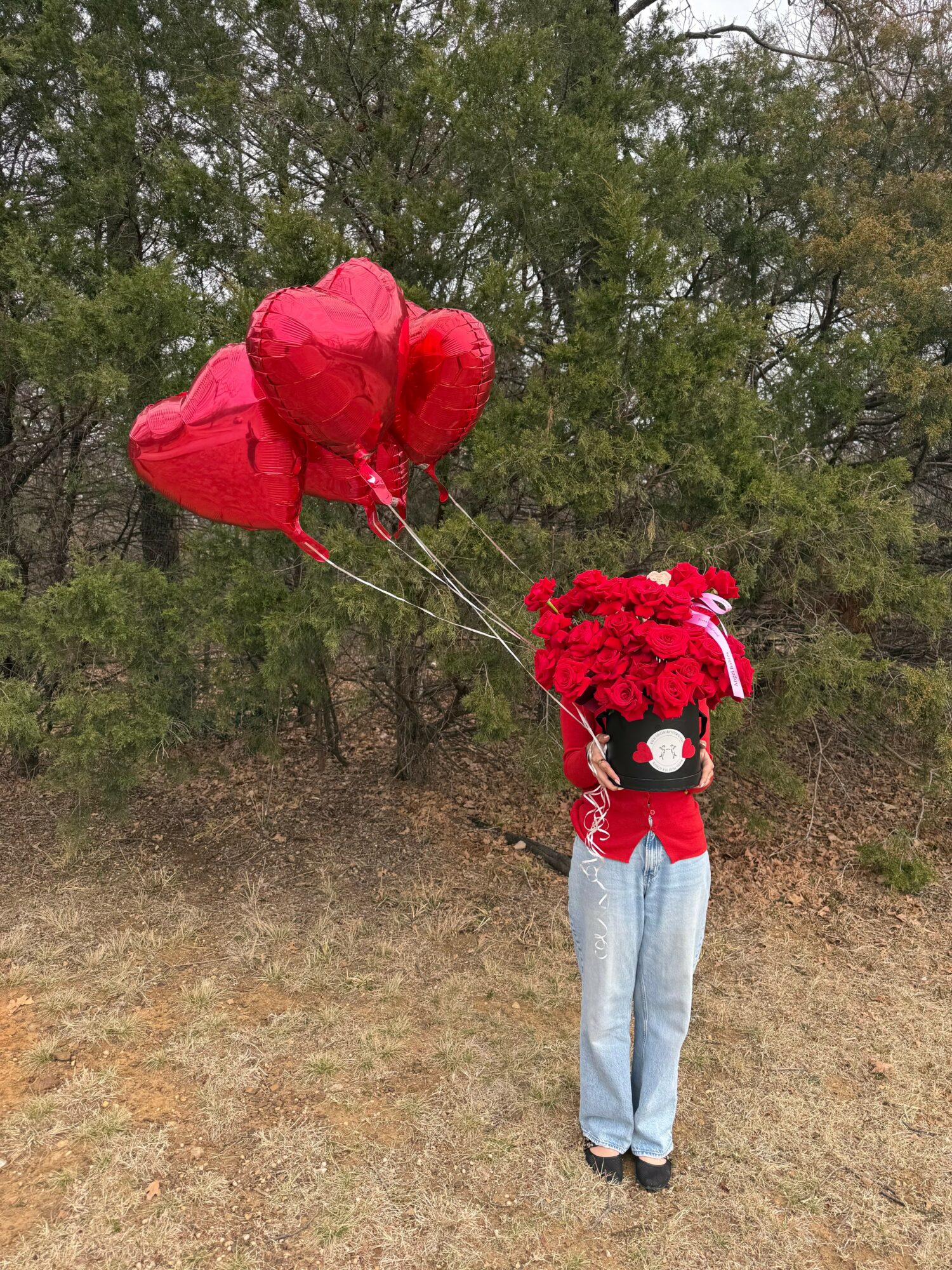 Person holding red heart-shaped balloons and a bouquet of red flowers outdoors, with trees in the background.