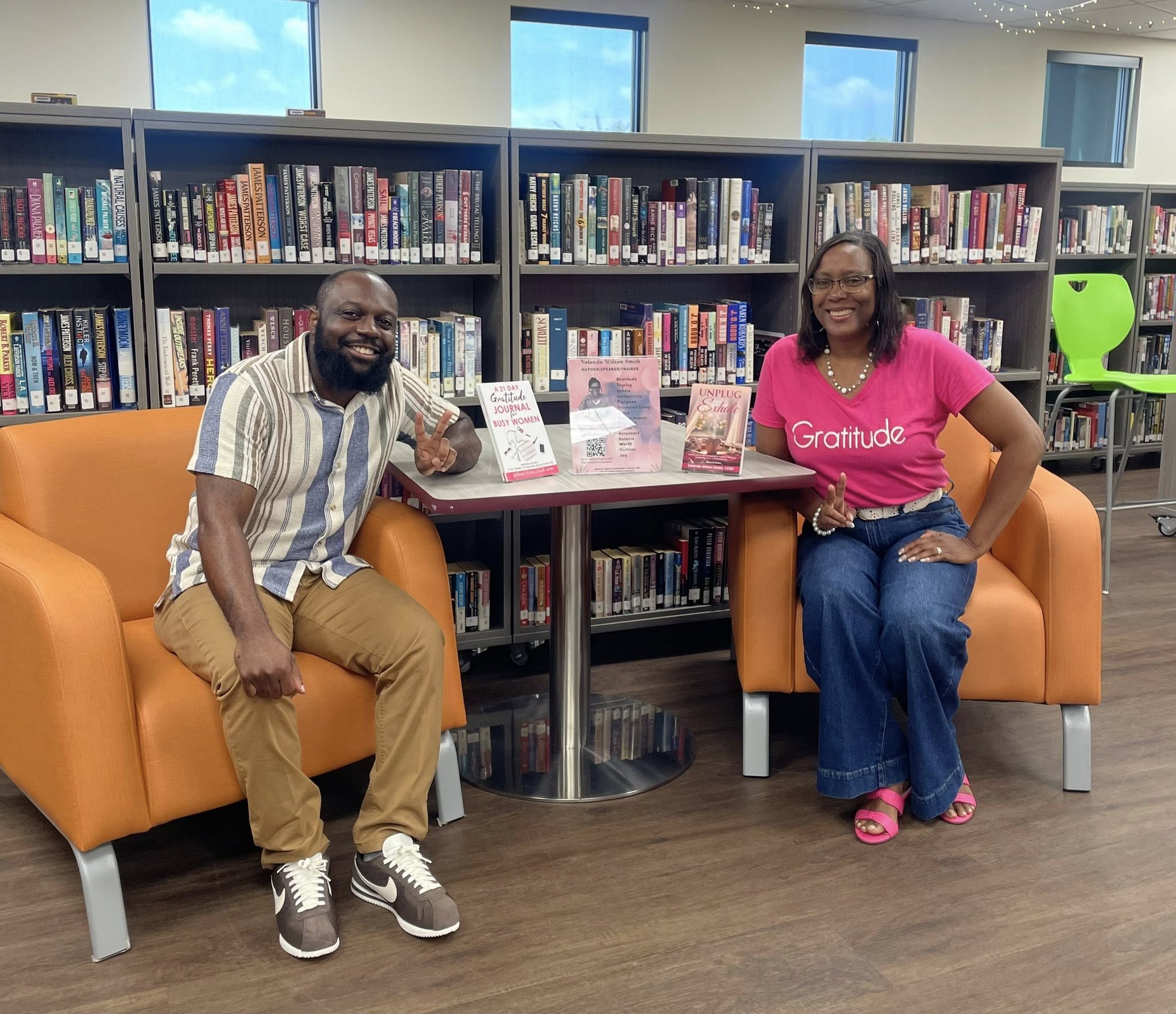 Two people sitting on orange chairs in a library, smiling, with bookshelves behind them and a table with books between them.