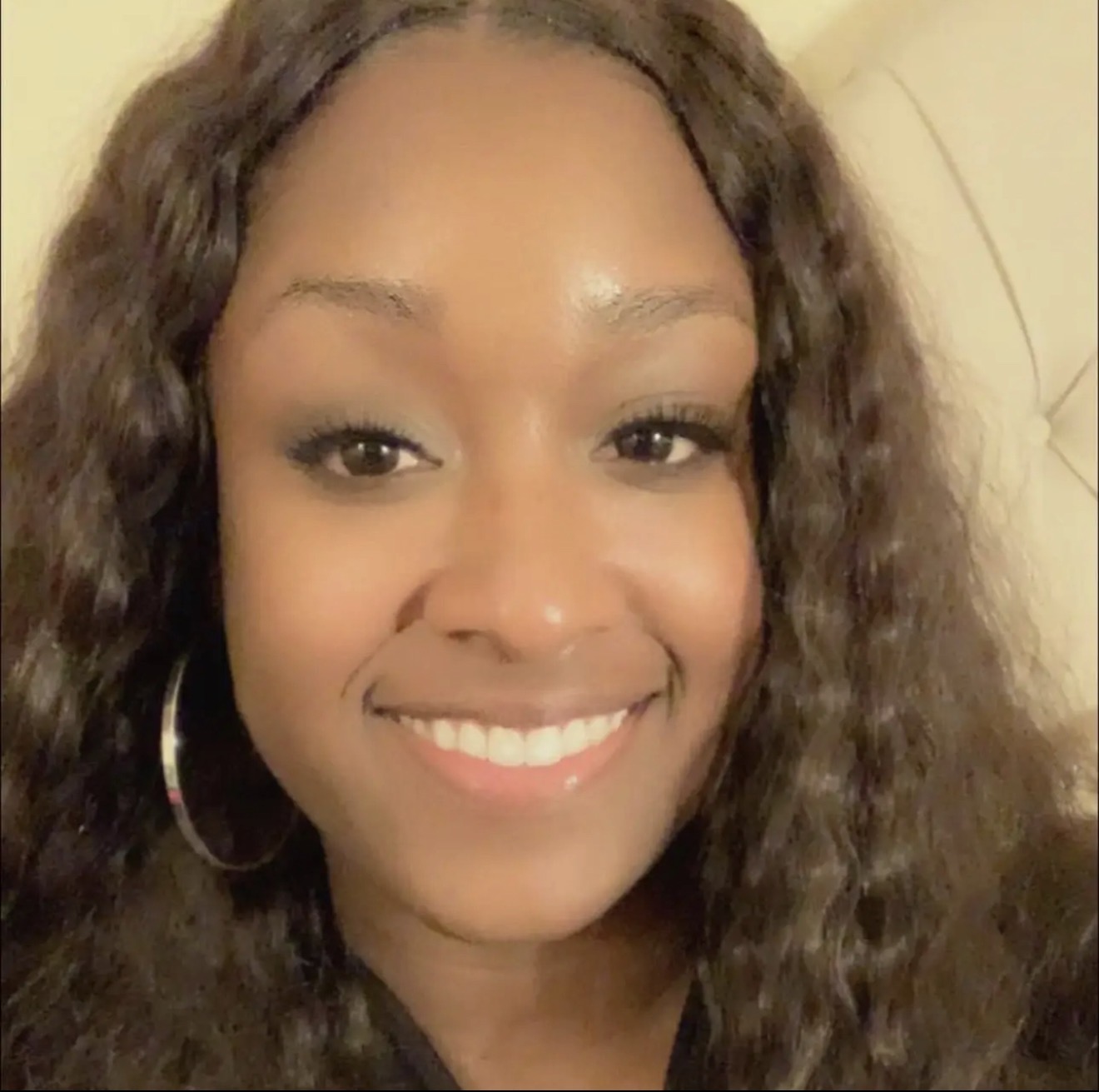 Close-up of smiling woman with curly hair, hoop earrings, and a light-colored background.
