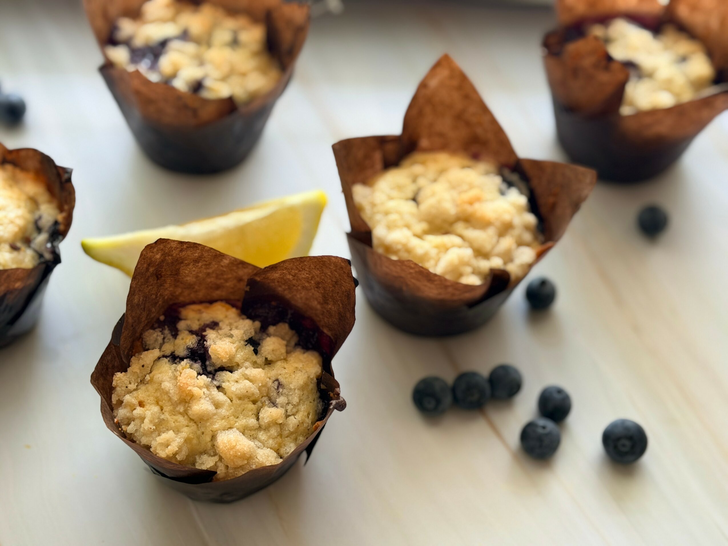 Four blueberry muffins with crumb topping, lemon wedge, and blueberries on a white surface.